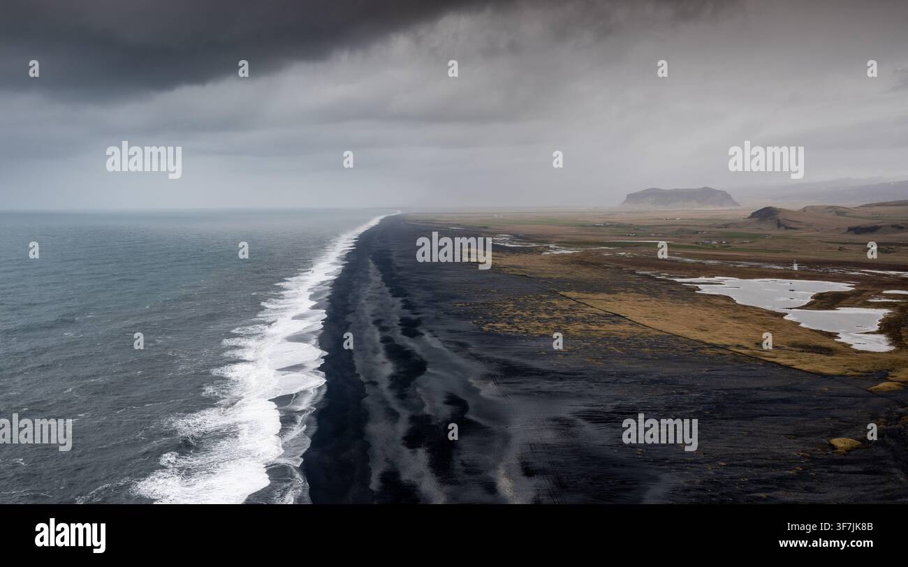 Spiaggia di sabbia nera di Reynisfjara dal punto panoramico della penisola di Dyrholaey. Paesaggio costiero spiaggia di sabbia vulcanica nera onde dell'oceano Atlantico cielo cupo Islanda ic Foto Stock