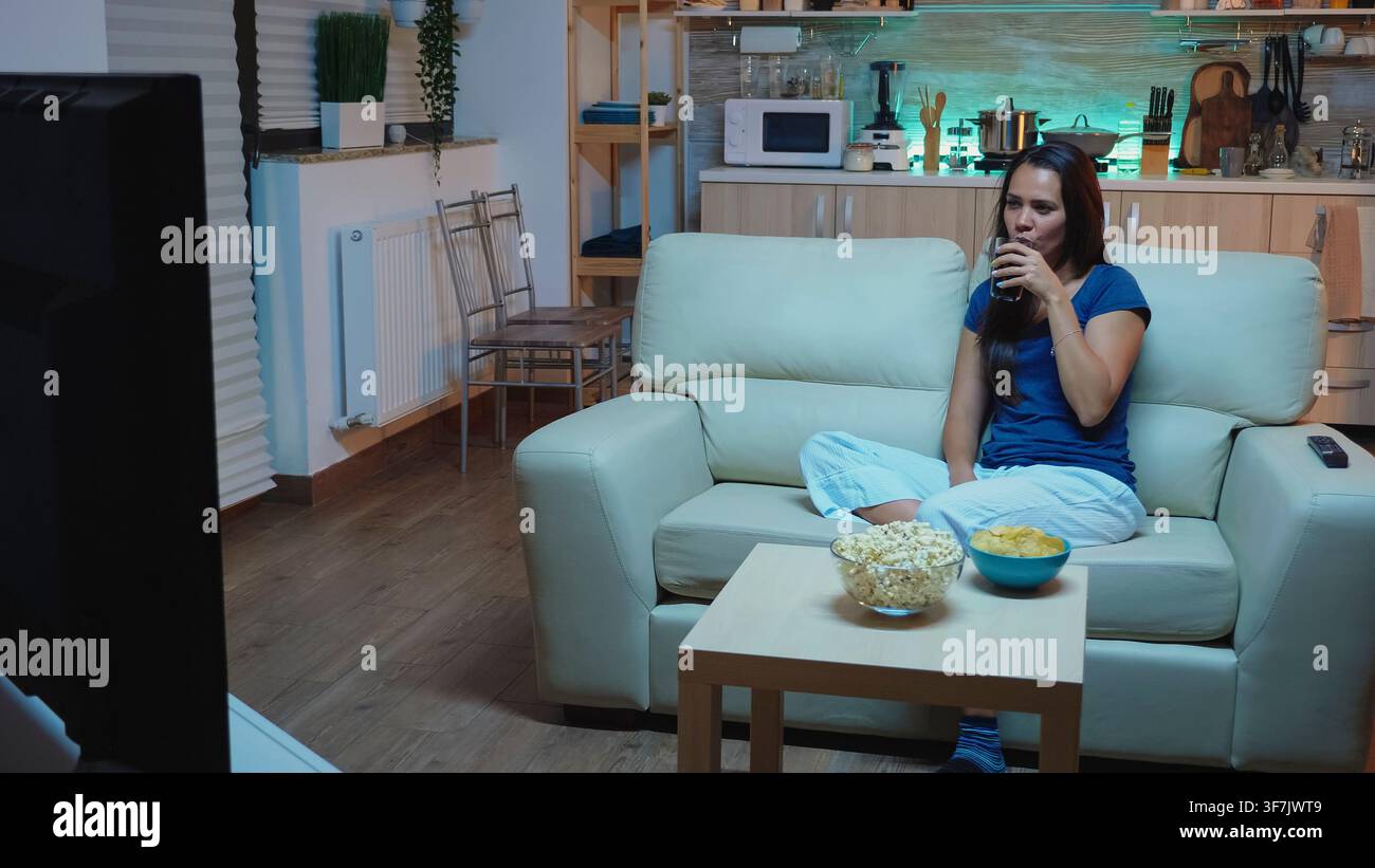 A casa, la donna ride di un programma televisivo divertente mentre gli spuntini rimangono a portata di mano durante il tempo libero serale allegro. Foto Stock