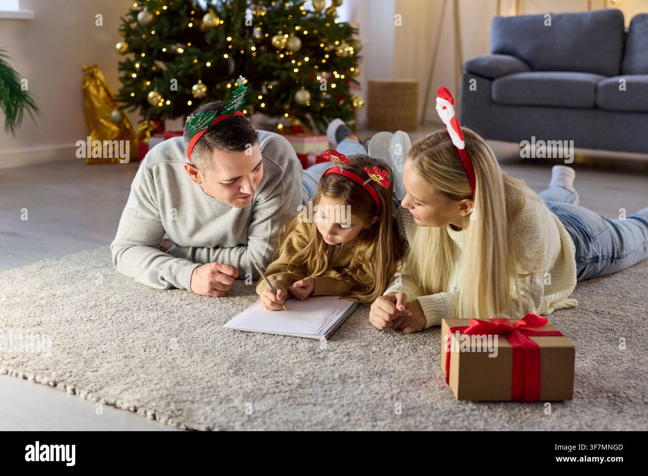 Famiglia felice che aiuta il bambino a scrivere una lettera di Natale sul pavimento vicino a un albero decorato Foto Stock