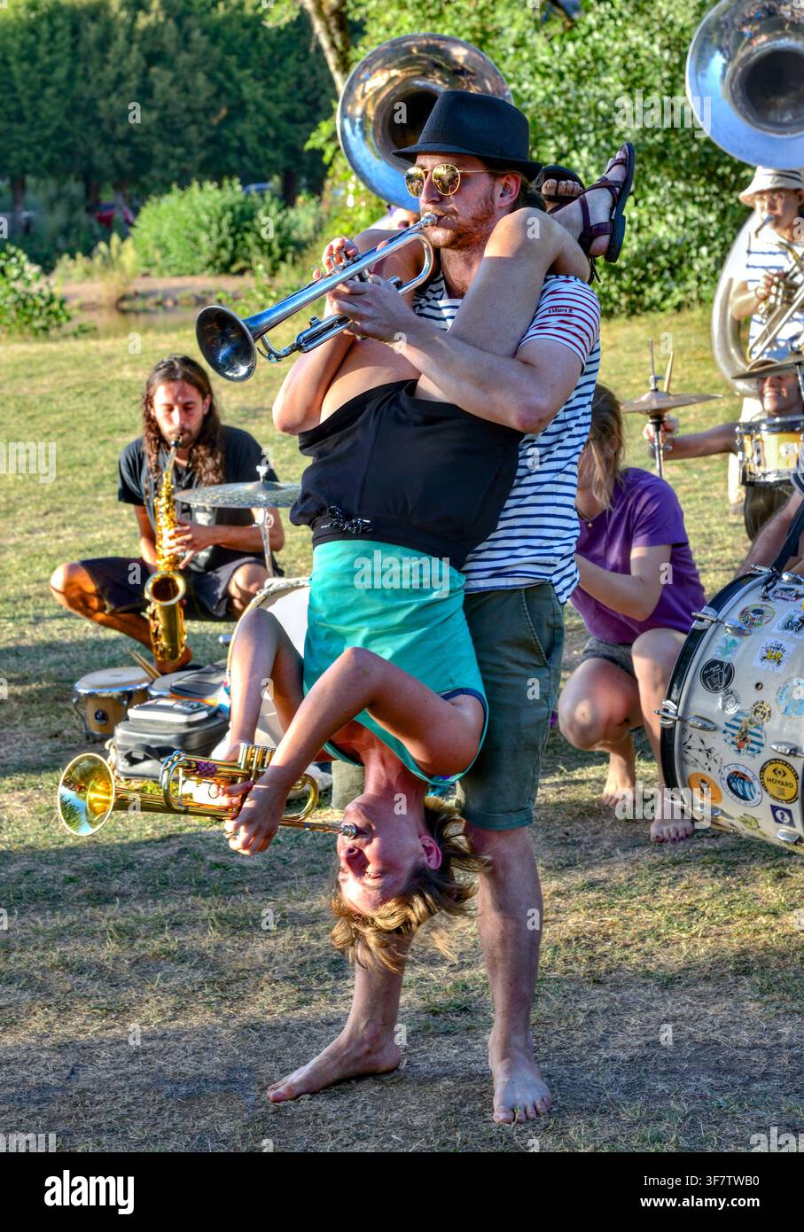 Uomo con donna appesa a testa in giù che suona trombe con la band Fanfan de Bordeaux in un concerto all'aperto - Bélâbre, Indre (36), Francia. Foto Stock