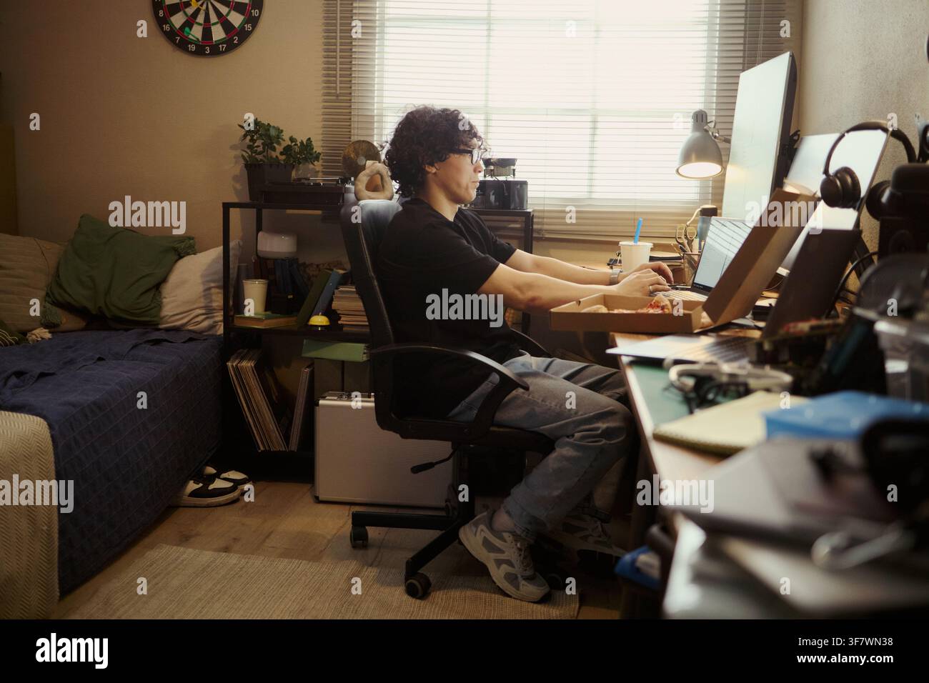 Giovane uomo asiatico che lavora al computer in camera da letto, digita sulla tastiera e mangia pizza. L'immagine rappresenta il lavoro a distanza, lo stile di vita freelance e il marketing degli alloggi per studenti Foto Stock