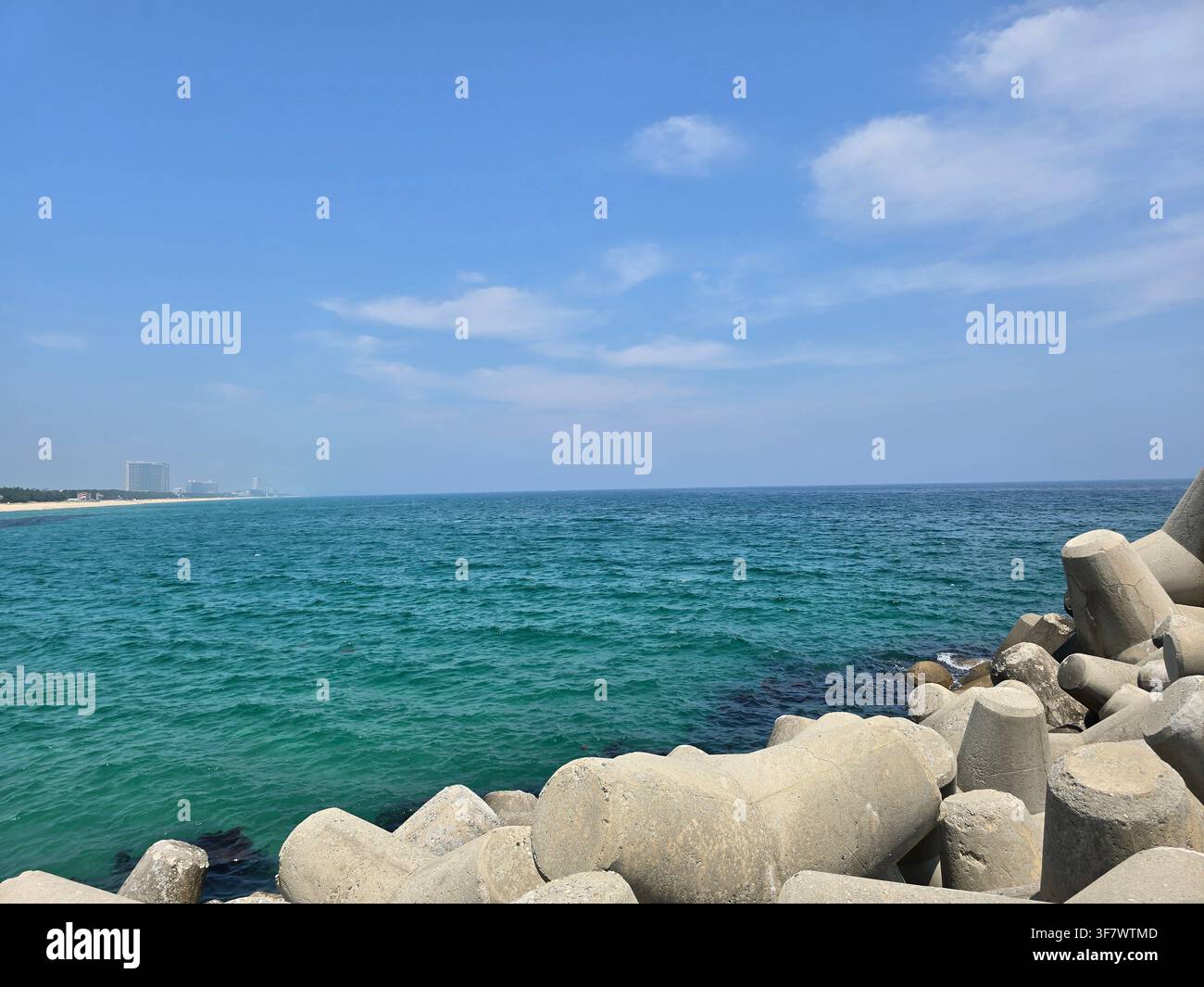 Blocchi frangiflutti in calcestruzzo a forma di tetrapode in primo piano con una vasta veduta del mare turchese e del cielo blu, la costa sabbiosa lontana visibile all'orizzonte - Immagine stock catturata con smartphone