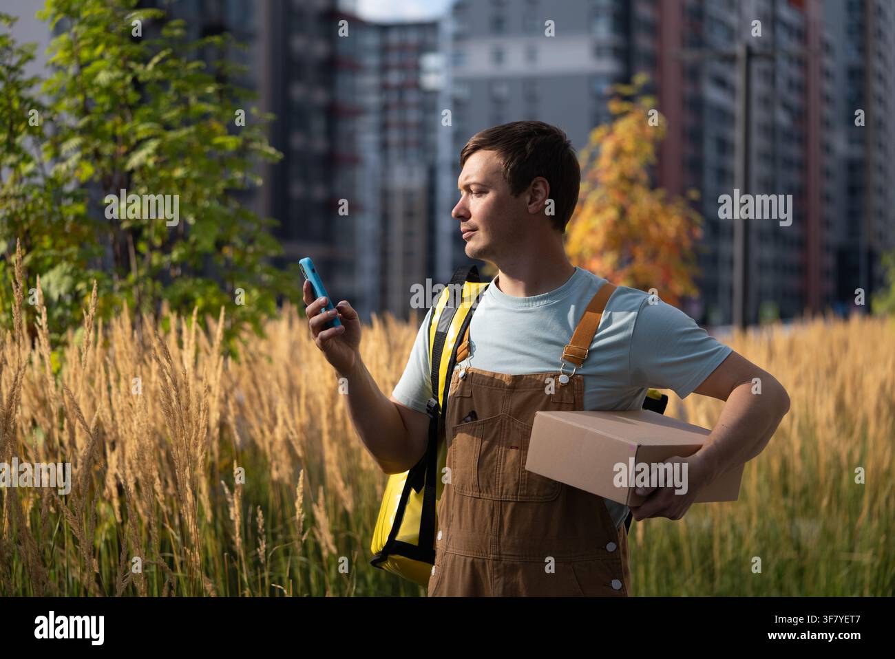 Corriere maschile che elabora un ordine su uno smartphone e trasporta un pacco di consegna, raffigurando i servizi di consegna urbani e il flusso di lavoro dinamico di un lavoratore dell'economia gig Foto Stock