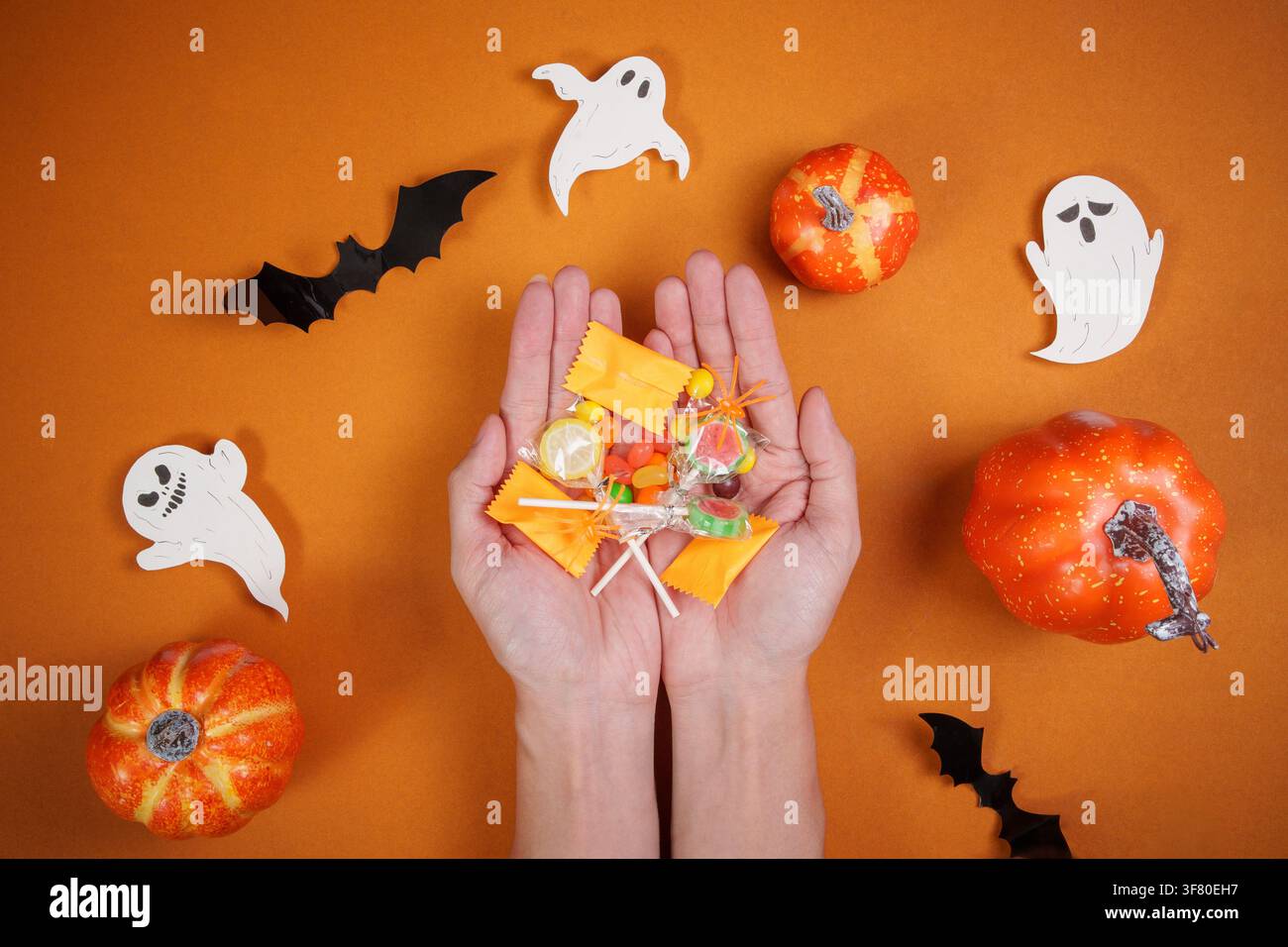 Mani che tengono una varietà di dolcetti di Halloween su uno sfondo arancione con zucche e fantasmi Foto Stock
