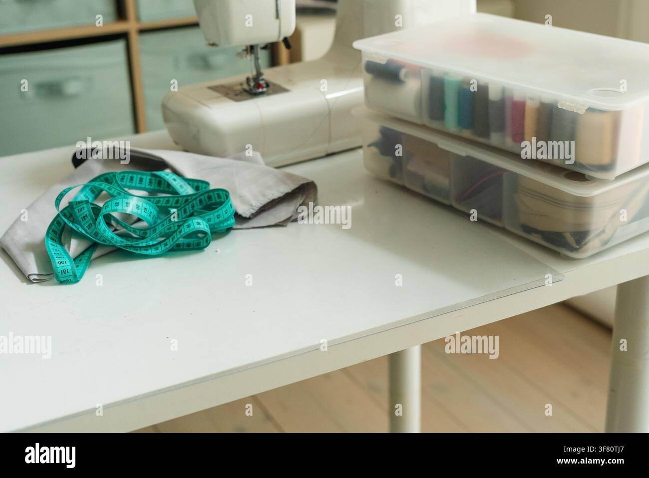 Una macchina da cucire su un tavolo bianco con tessuto grigio e un metro per la preparazione. Strumento per il fai-da-te pronto per cucire il tessuto con calma e concentrazione organizzata in uno studio domestico con luce naturale. Foto Stock