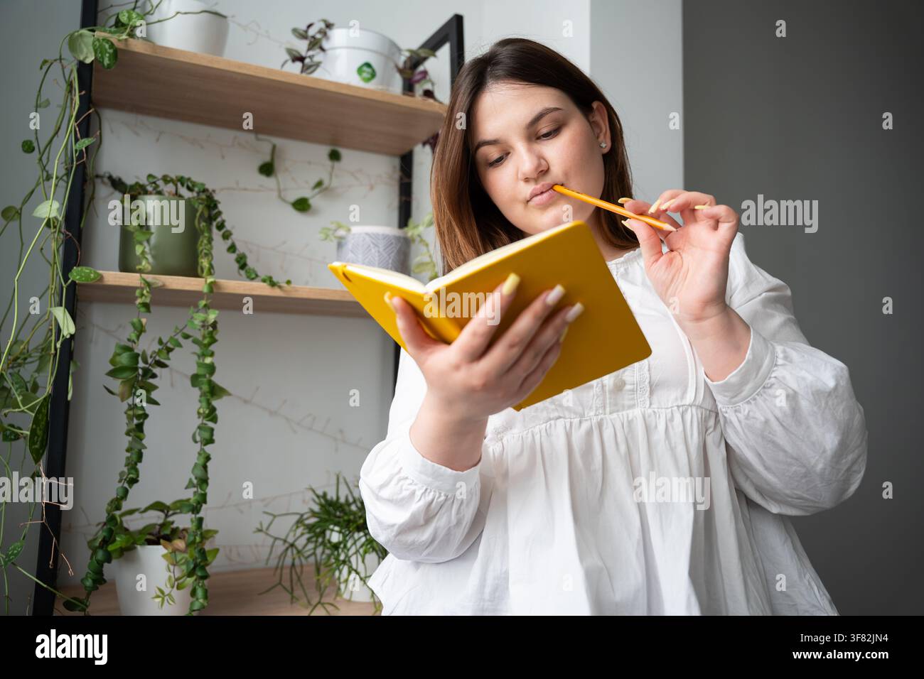 Giovane donna che tiene pensierosamente un quaderno e una matita in un ambiente domestico. Studio a casa, pianificazione personale e routine di giornalismo. Foto Stock