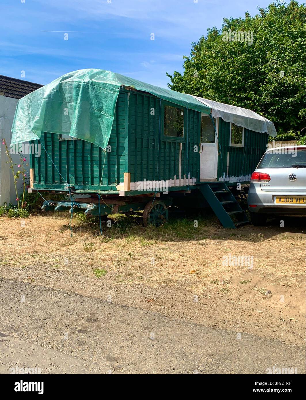 Carovana Gypsy in legno verde restaurata sotto i cieli estivi, parcheggiata accanto a un'auto su un terreno asciutto in Inghilterra con una copertura sopra - Immagine stock catturata con smartphone