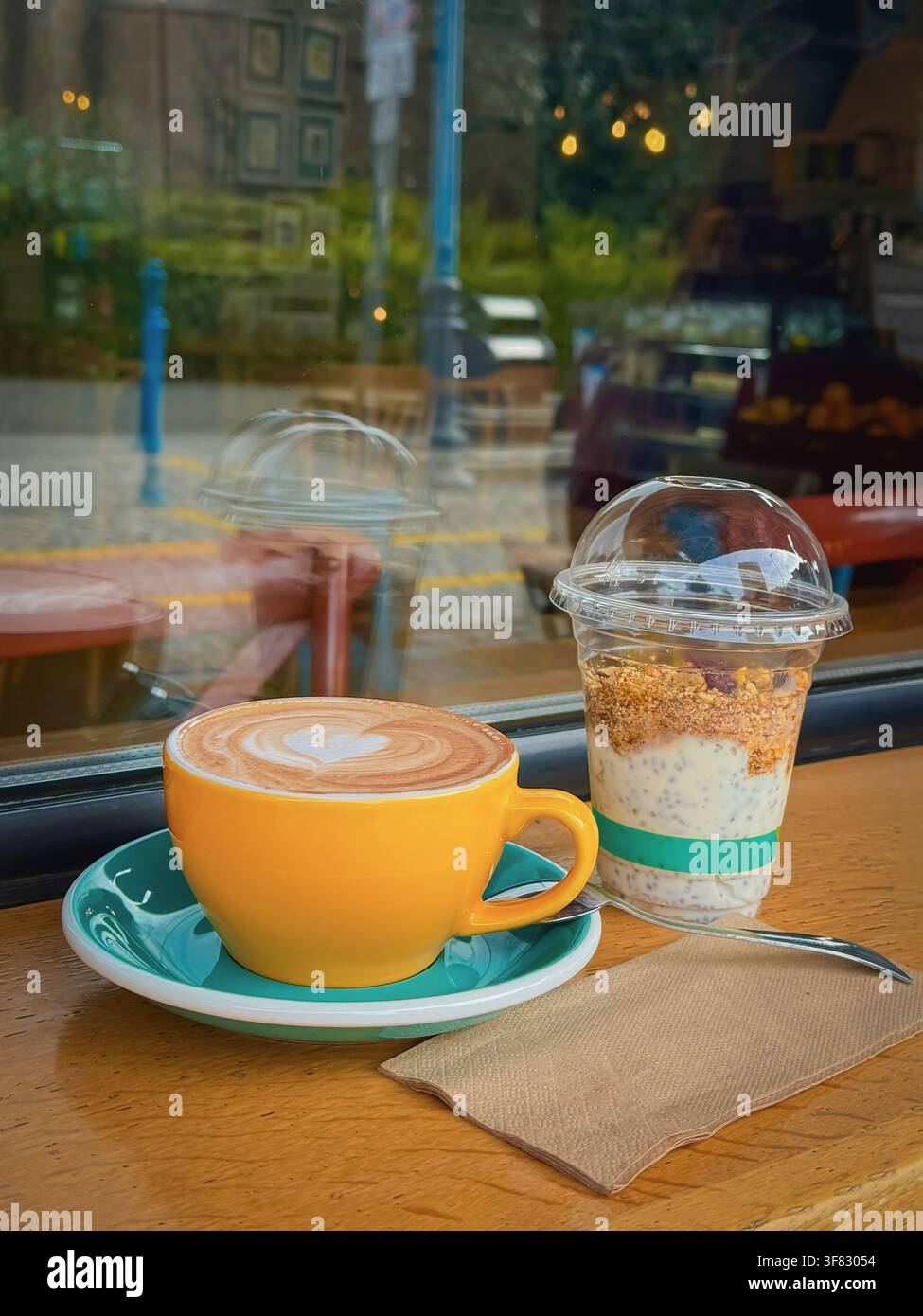 Tazza gialla di latte fresco con latte art a forma di cuore accanto a un dessert cremoso al chia su un tavolo di legno del caffè, riflessi nel vetro della finestra. Caffè luminoso Foto Stock