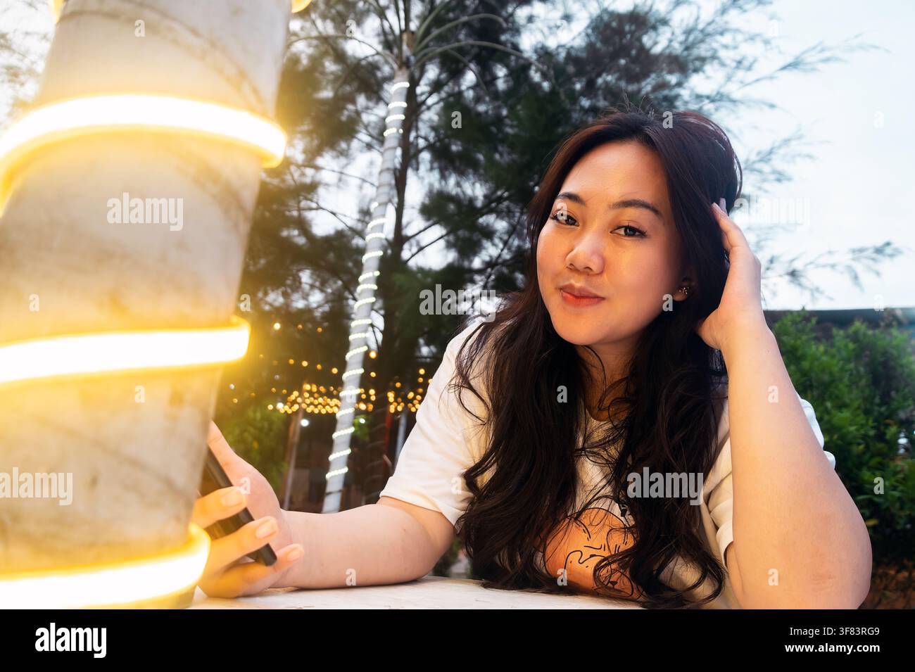 giovane donna asiatica che guarda la fotocamera con un sorriso sottile mentre è seduta all'aperto la sera, tenendo uno smartphone vicino a un'illuminazione decorativa calda Foto Stock
