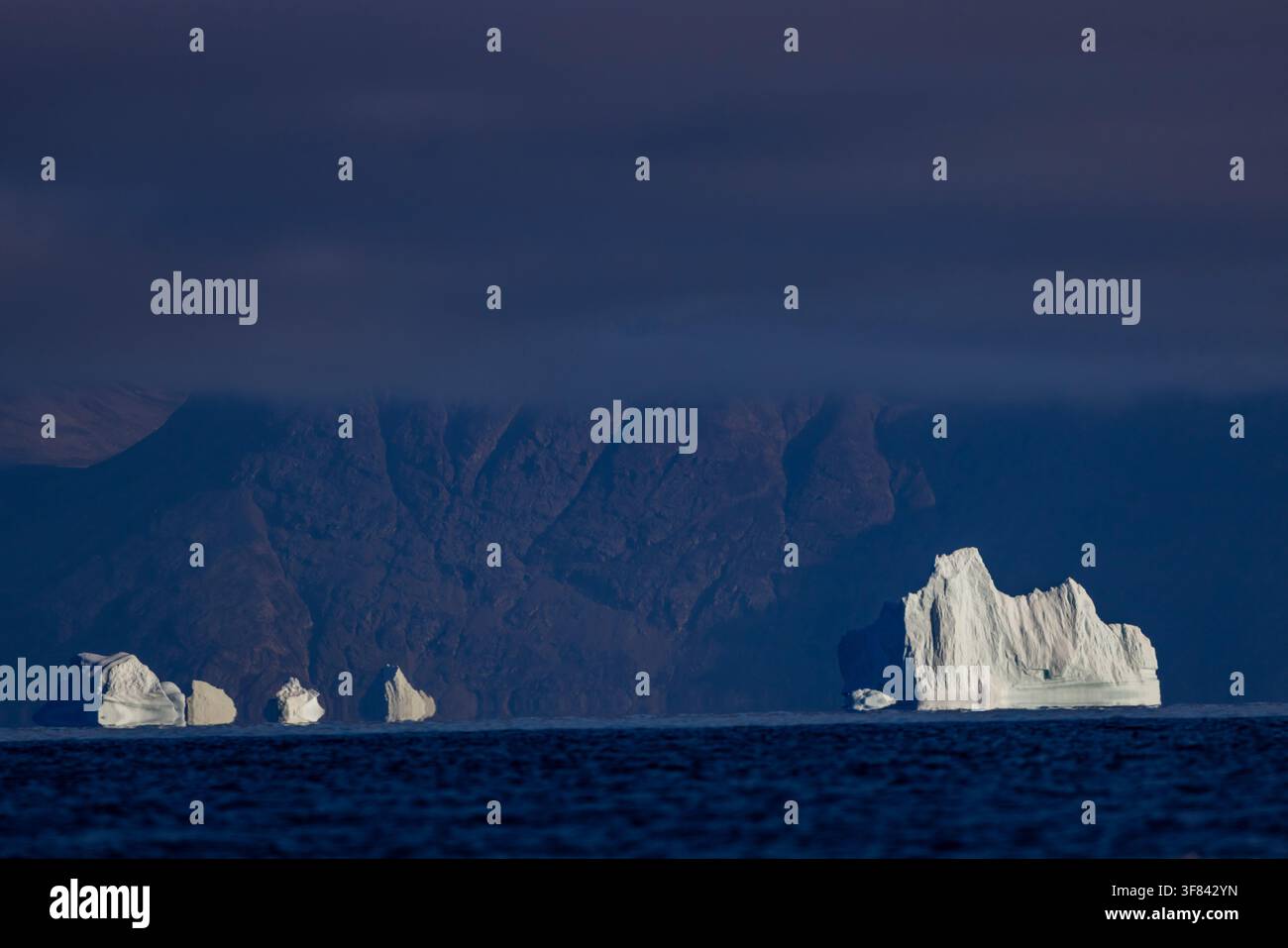 Gli iceberg brillano sotto nuvole scure con uno sfondo di isole rocciose nelle acque artiche della Groenlandia Foto Stock