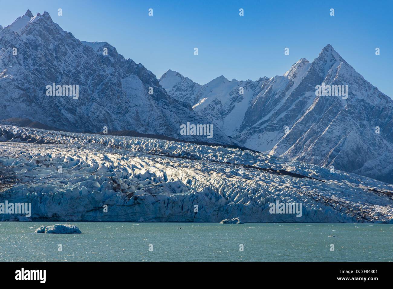 Picchi frastagliati si ergono sopra una fronte glaciale con iceberg blu che galleggiano nelle acque artiche vicino a Blomsterbugten in Groenlandia Foto Stock