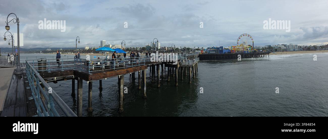 Le persone camminano sul molo che si affaccia sull'oceano con giostre e attrazioni in lontananza durante una giornata nuvolosa in California Foto Stock