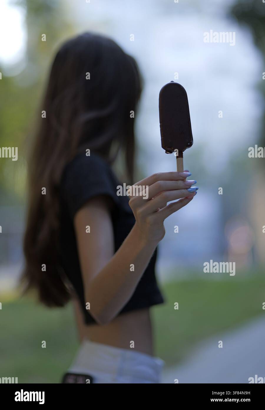 Giovane donna che tiene una barretta di gelato al cioccolato che si scioglie su uno stecco, godendosi un dolce refrigerante all'aperto con il caldo, simboleggiando il tempo libero estivo Foto Stock