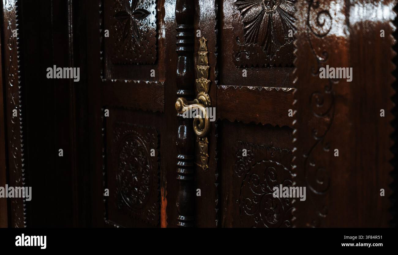 Maniglia decorativa in ottone montata su una porta in legno intagliata in modo intricato. Artigianato tradizionale e dettagli architettonici storici. Foto Stock
