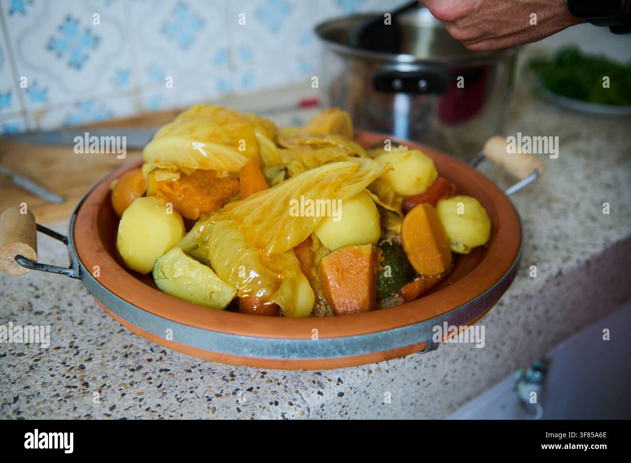 Couscous marocchino tradizionale con verdure al vapore e foglie di cavolo su un piatto di terracotta, presentato su un bancone della cucina di casa pronto per essere servito, wa Foto Stock