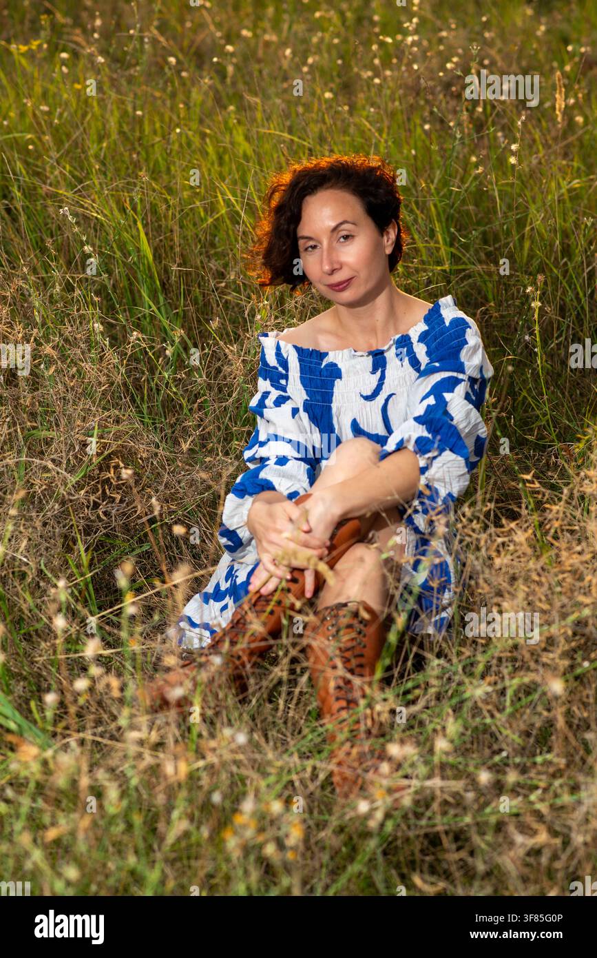 Donna sorridente seduta in un prato illuminato dal sole, indossa un vestito blu a spalle scoperte e stivali con lacci marroni, ritratto dello stile di vita rilassato. Foto Stock