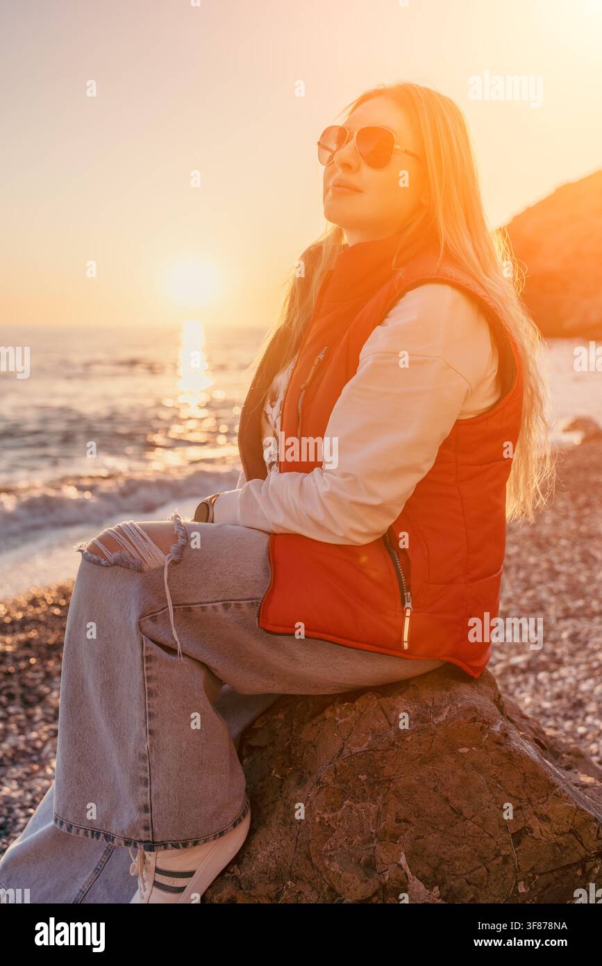 Donna seduta su una scogliera costiera all'ora dorata durante il tramonto, godendosi la luce calda della sera e le onde pacifiche dell'oceano Foto Stock