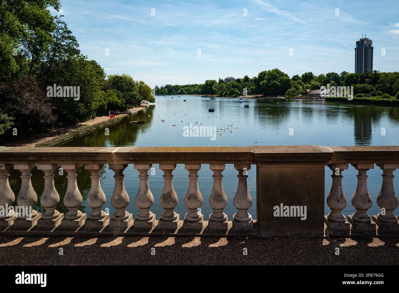 Vista sulla Serpentine a Hyde Park, Londra, incorniciata da una balaustra in pietra con alberi, barche e una torre sotto un cielo azzurro. Foto Stock