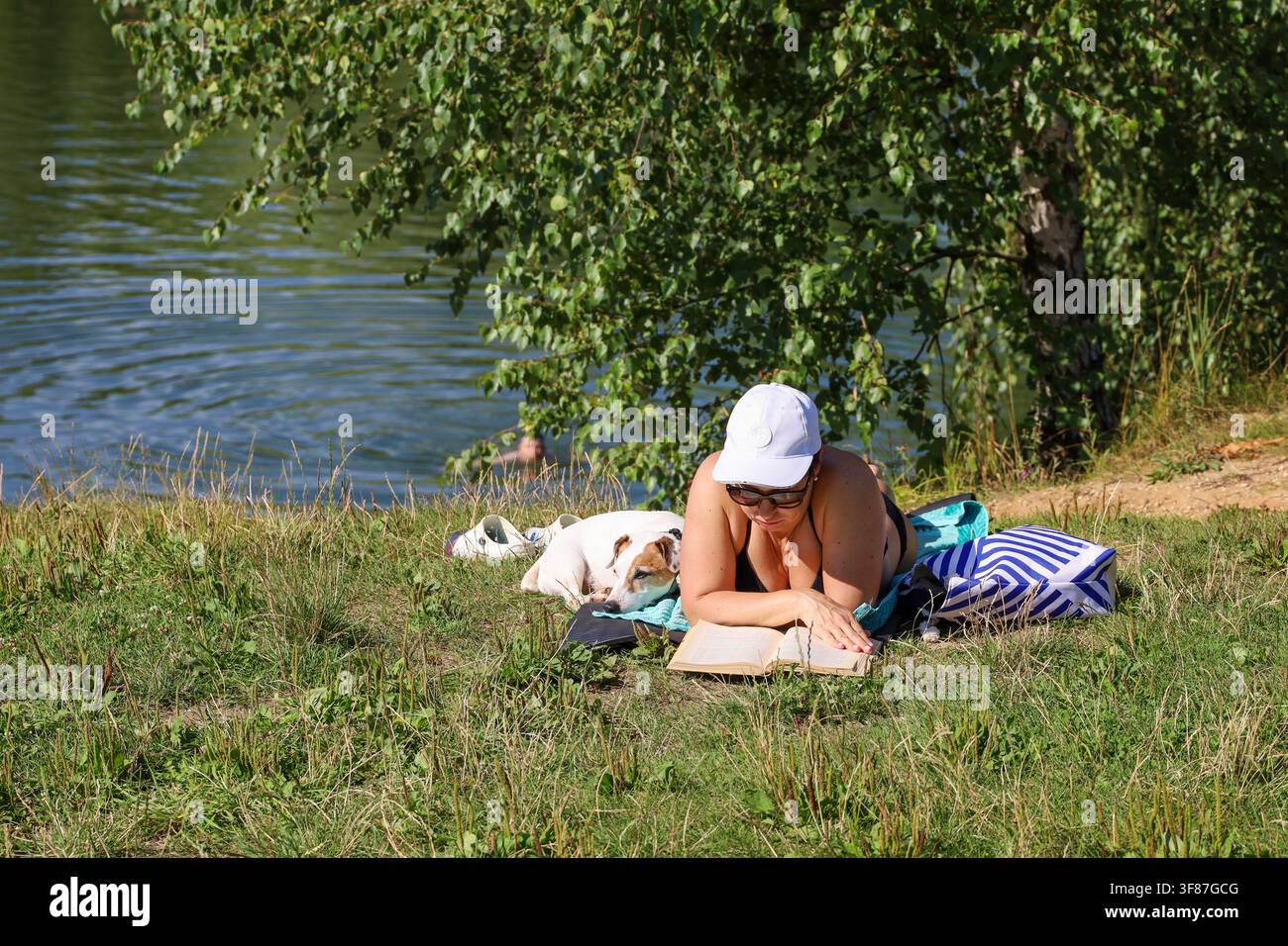 Donna legge un libro cartaceo sdraiata con il suo cane sulla spiaggia del fiume, tempo libero estivo Foto Stock