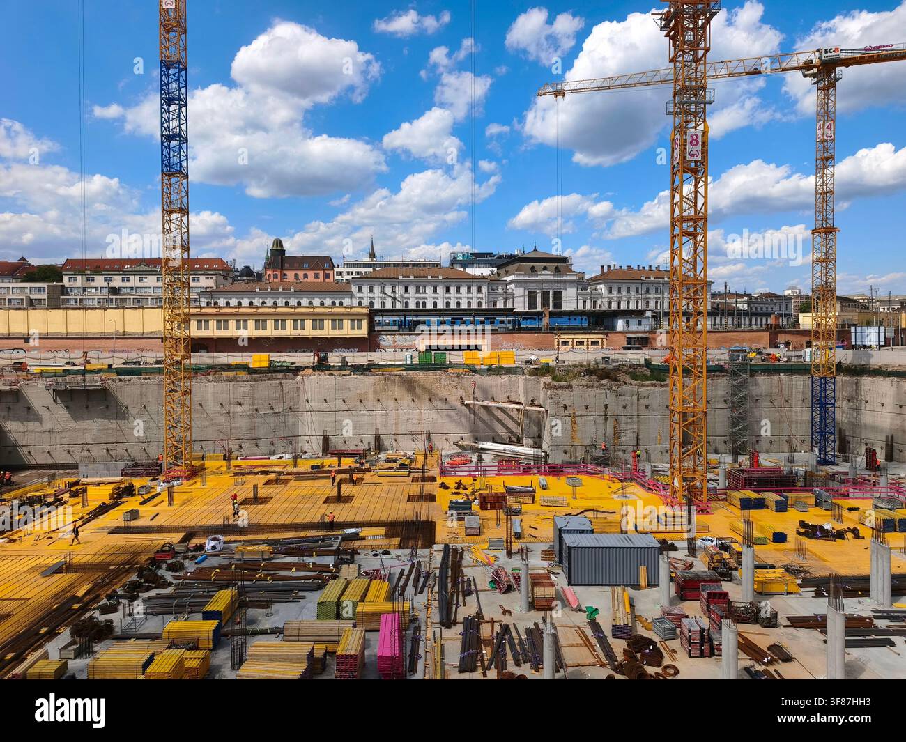 Grande fossa di cantiere con gru gialle, telaio di fondazione e operai con l'edificio principale della stazione ferroviaria e un treno sullo sfondo - Immagine stock catturata con smartphone