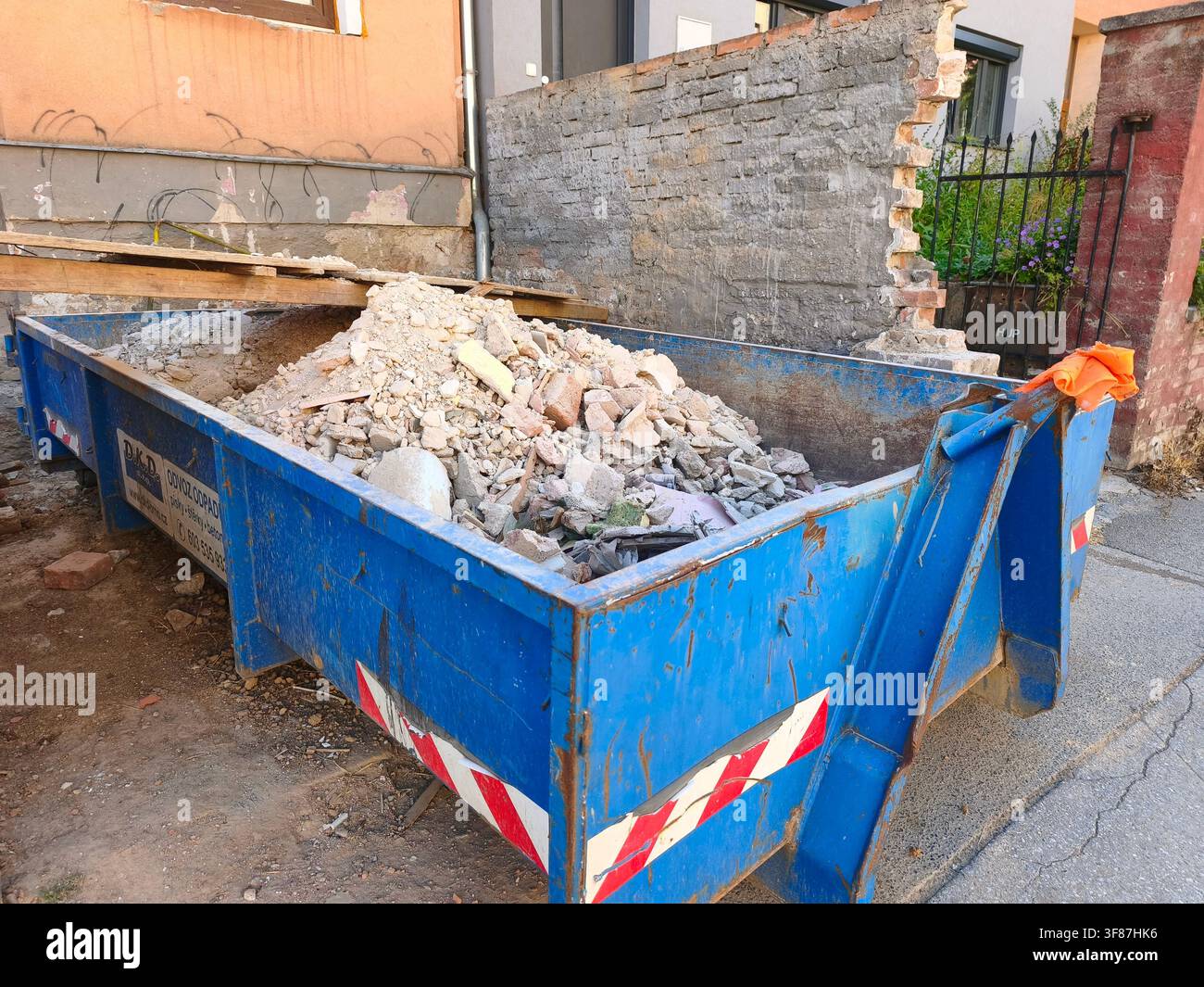 Grande contenitore industriale blu pieno di detriti da costruzione e macerie di un muro di mattoni demolito accanto a una casa - Immagine stock catturata con smartphone