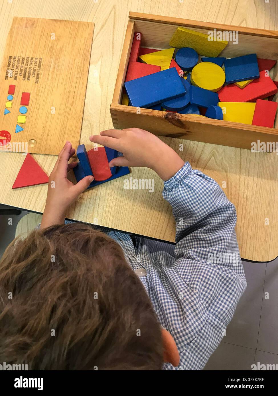 Vista dall'alto di un bambino in grembiule scolastico che costruisce e gioca con blocchi di forme geometriche in legno colorati a una scrivania di legno in un ambiente scolastico Foto Stock