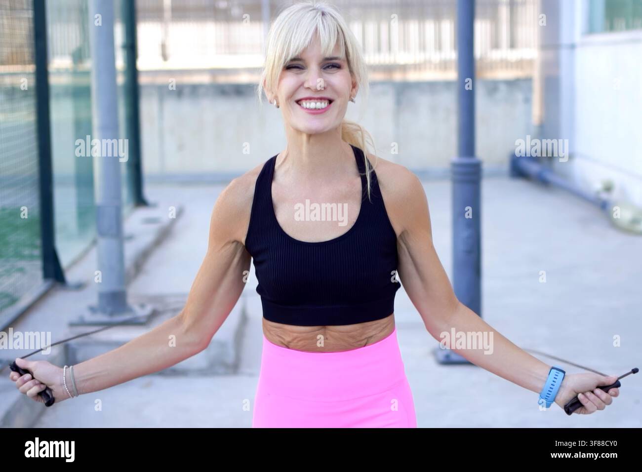 Giovane donna sorridente che salta la corda all'aperto su un campo illuminato dal sole, indossa un reggiseno sportivo nero, leggings rosa acceso, capelli biondi corti raccolti, smartwatch al polso Foto Stock