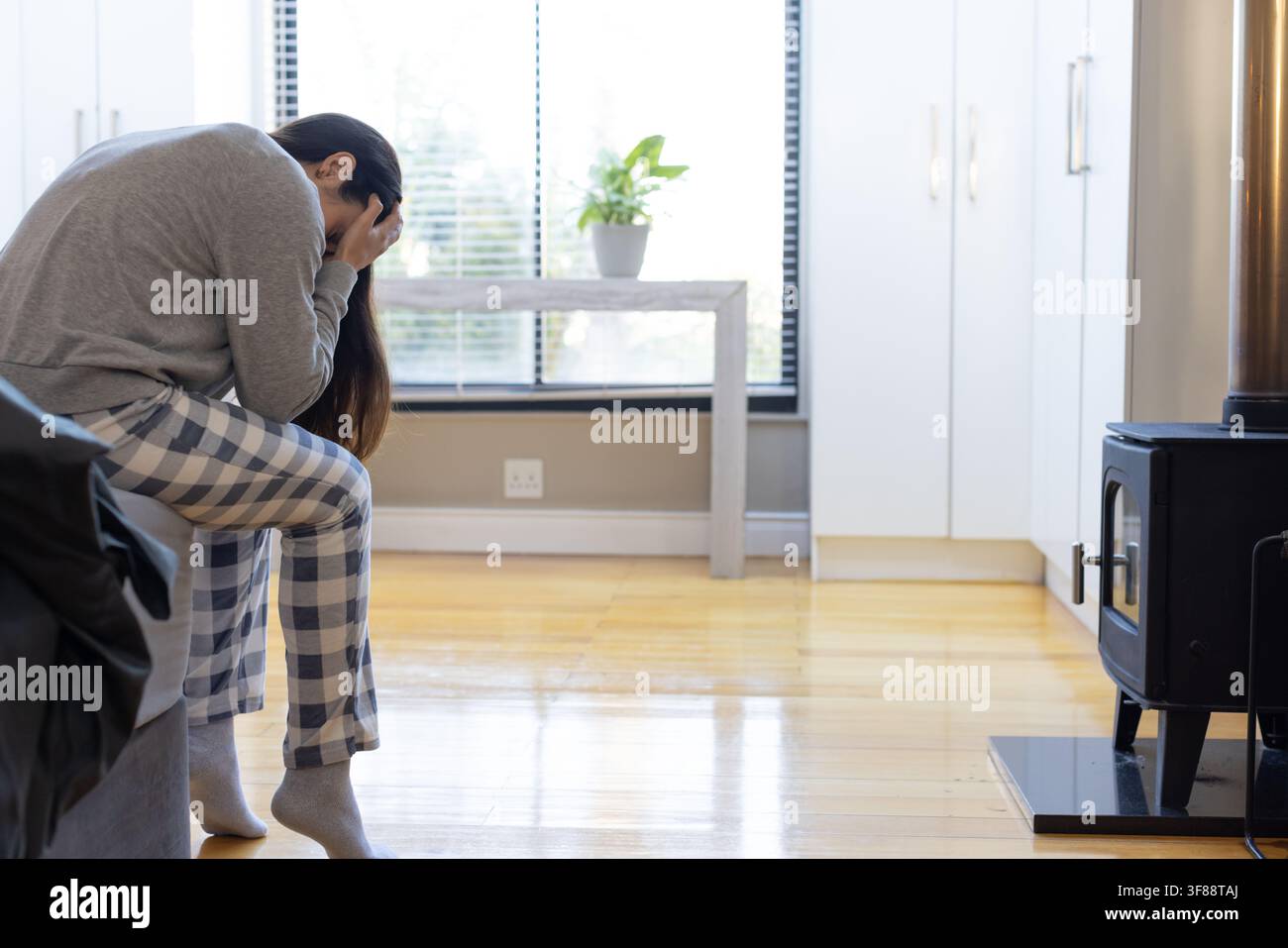 Donna seduta sul divano con pantaloni da pigiama a quadri, tiene il viso tra le mani a casa, spazio di copia. Solitaria, contemplativa, accogliente, abbigliamento da relax, stufa a legna, bli Foto Stock