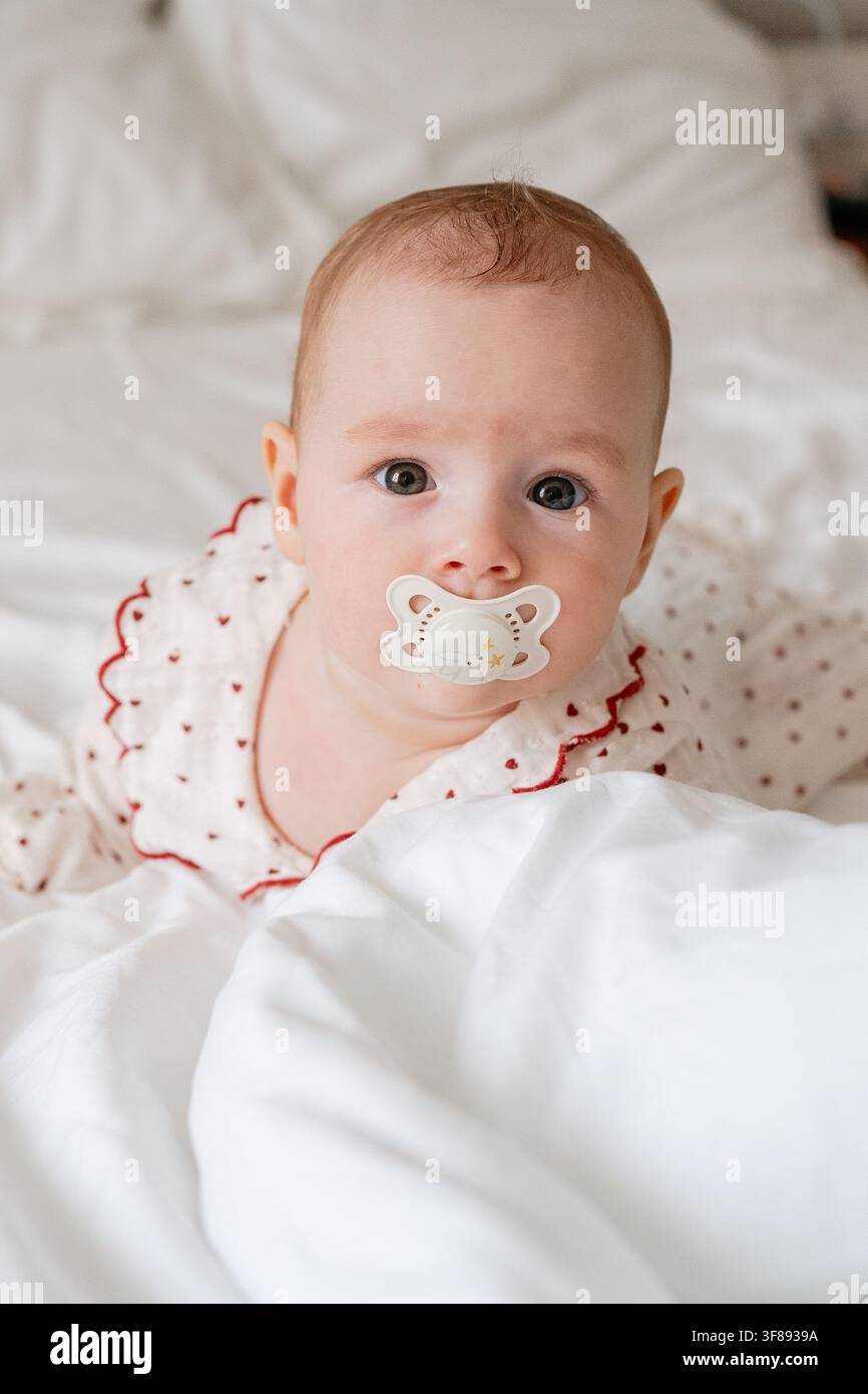 un bambino che guarda la telecamera con un ciuccio in bocca, emanando calma e soddisfazione in un ambiente accogliente da camera da letto, catturato in un primo piano con Foto Stock