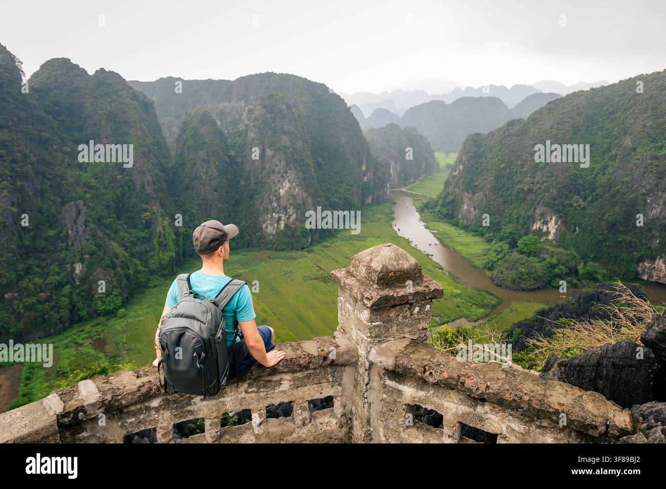 Vista posteriore di un turista con zaino seduto su un punto panoramico, che gode di una vista panoramica scenografica sui campi di riso e sul fiume circondati da montagne carsiche di calcare a N Foto Stock
