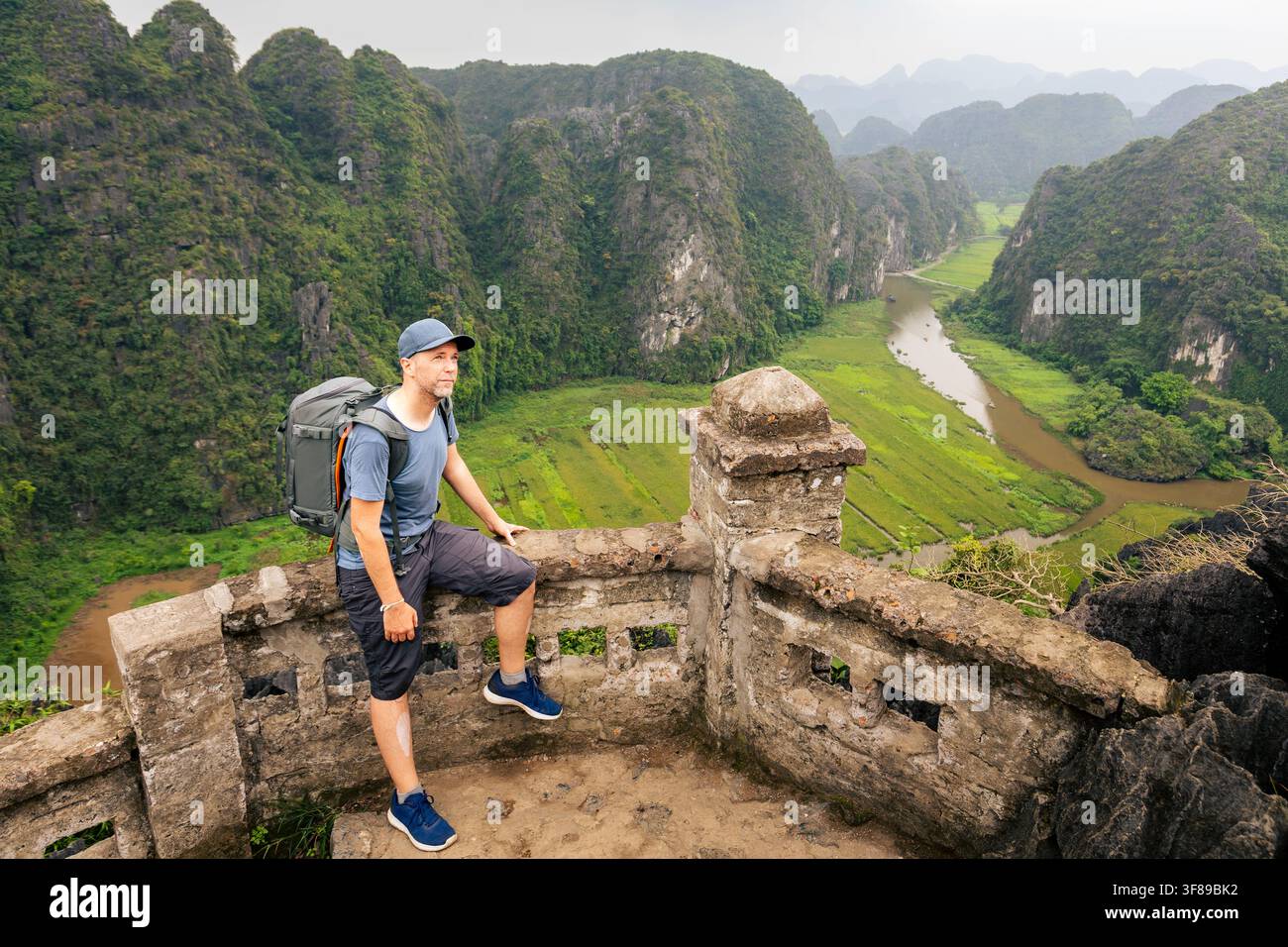 Turista con zaino che fa una pausa per riposare su una scogliera durante un'escursione in una giornata calda tra le montagne carsiche di calcare a Ninh Binh, Vietnam. Campi di riso, ventoso Foto Stock