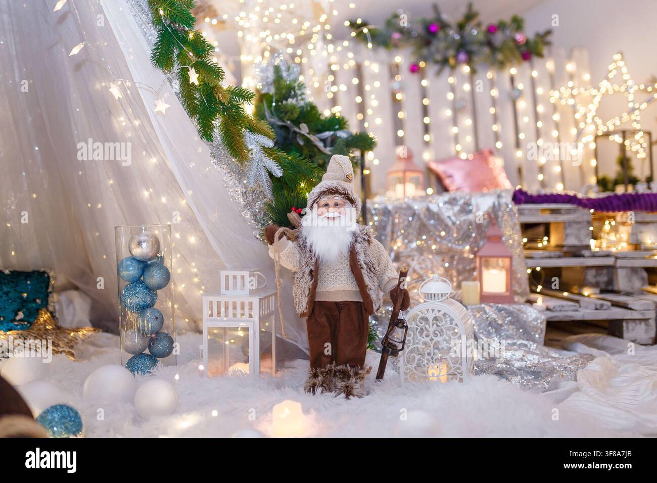 Bambola di Babbo Natale vestita di marrone in piedi su una lanterna decorativa bianca. Decorazione di Capodanno, luci scintillanti natalizie e bokeh su sfondo sfocato Foto Stock