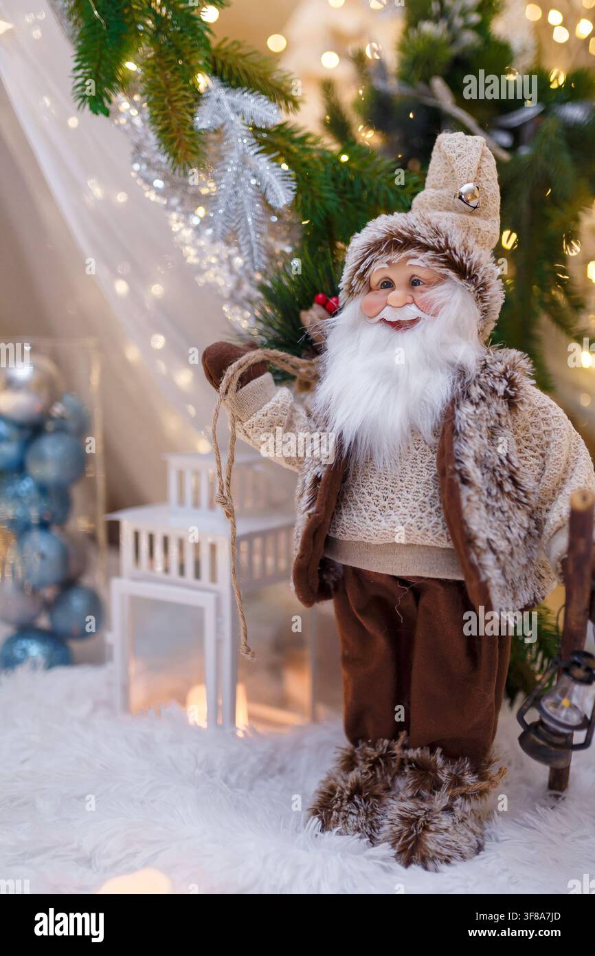 Giocattolo di Babbo Natale con vestiti marroni in piedi su una lanterna bianca. Rami di abete e decorazioni natalizie su sfondo sfocato con luci calde a ghirlanda Foto Stock