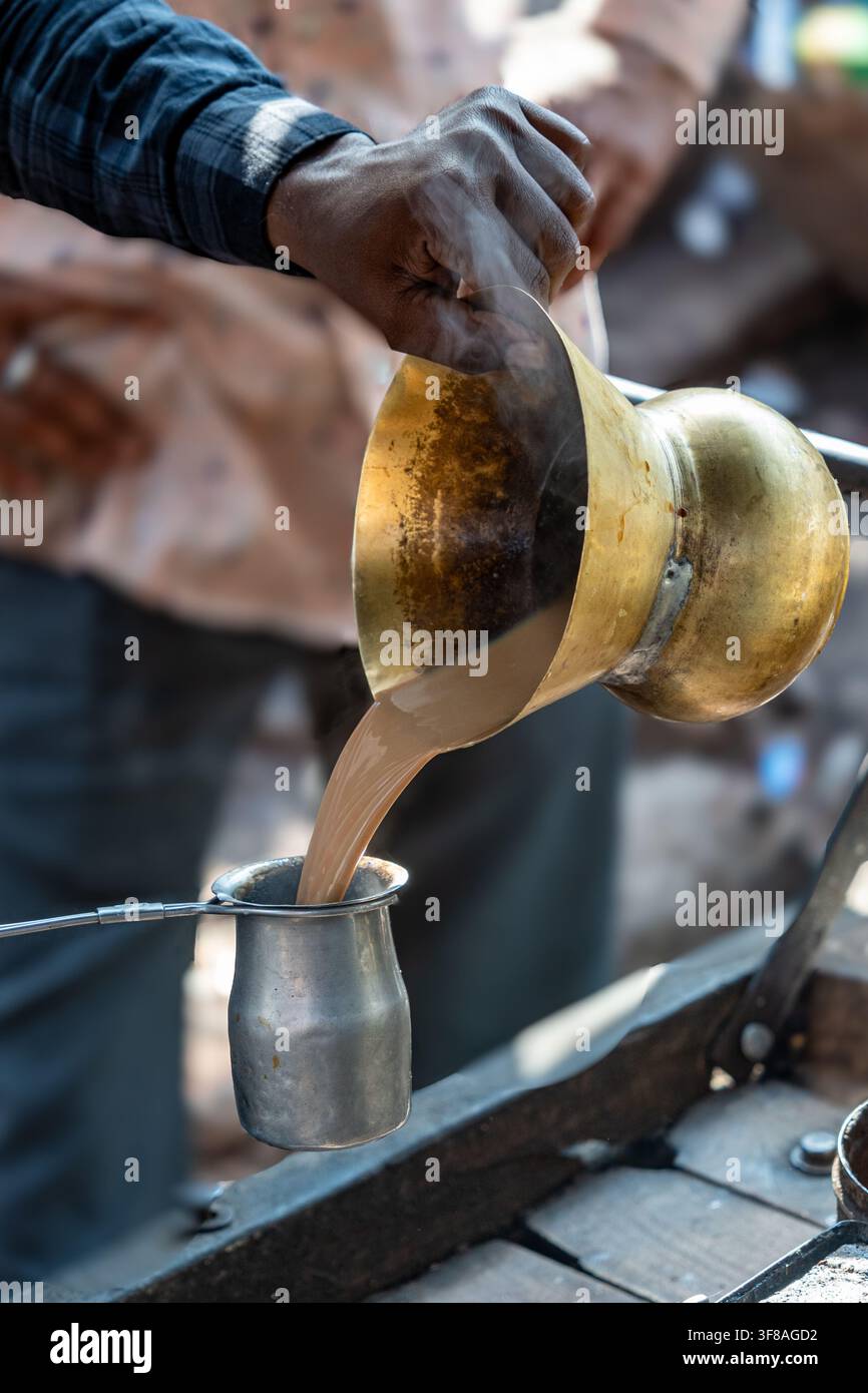 India. Rajasthan. Bundi. Preparazione del chai (tè indiano). Primo piano di un venditore ambulante che versa del chai fumante, preparato in una teiera di ottone Foto Stock