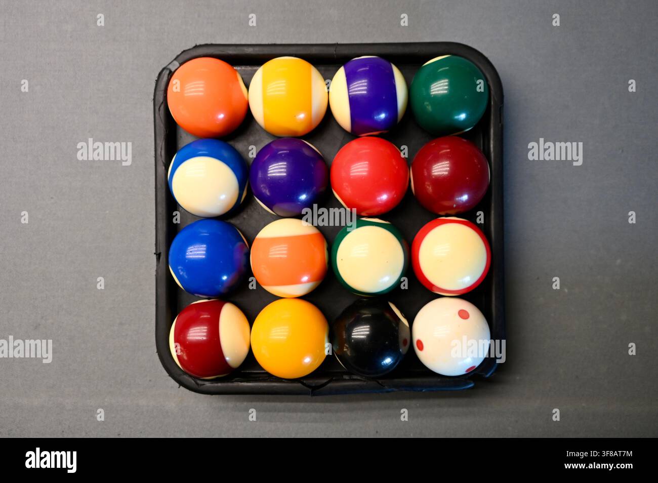 Fotografia commerciale a vista dall'alto di un set completo di palle da biliardo e pool colorate, comprese palle piene, a strisce e da cue, disposte ordinatamente all'interno di un vassoio quadrato nero su una superficie grigia. Foto Stock