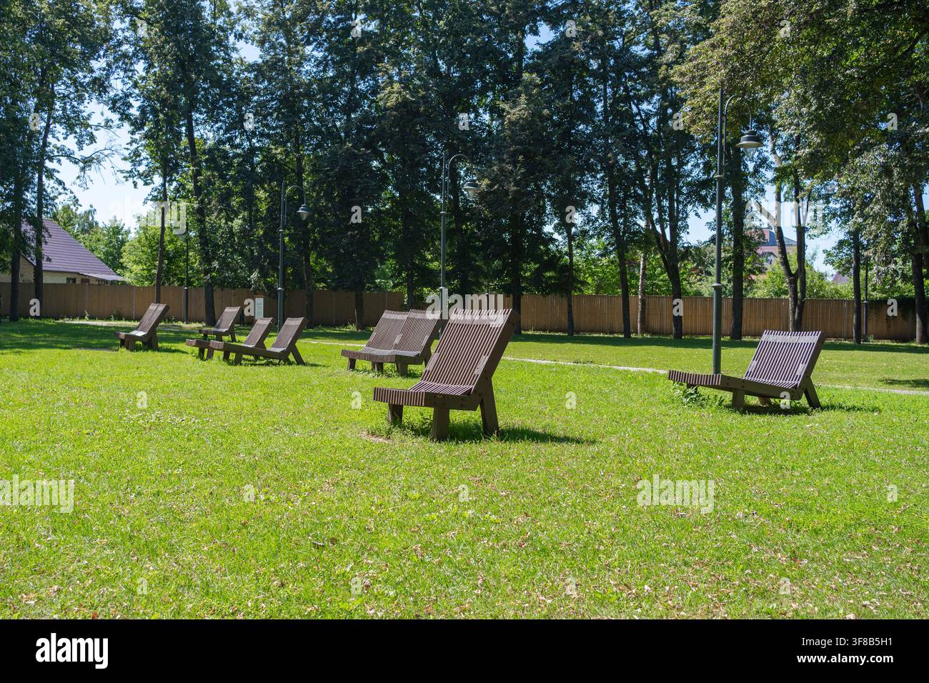 Area di riposo pubblica in un parco cittadino con sedie a sdraio ergonomiche in legno su erba verde circondata da alberi. Ideale per le vacanze estive, comfort urbano Foto Stock