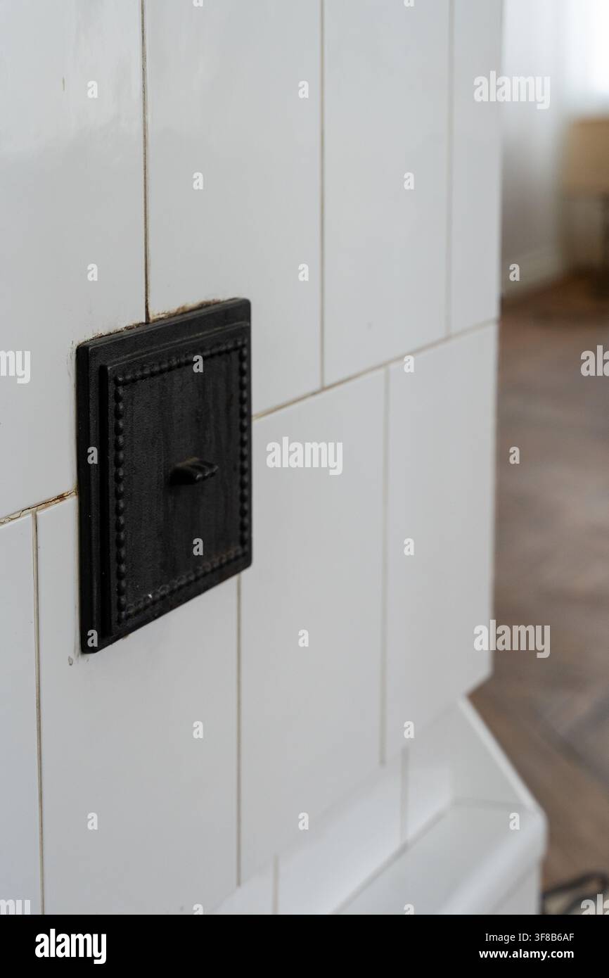 Prima di un tradizionale stufa in muratura con piastrelle bianche che mostra una porta di fuliggine in ghisa nera e una manopola del diaframma in metallo. Foto Stock