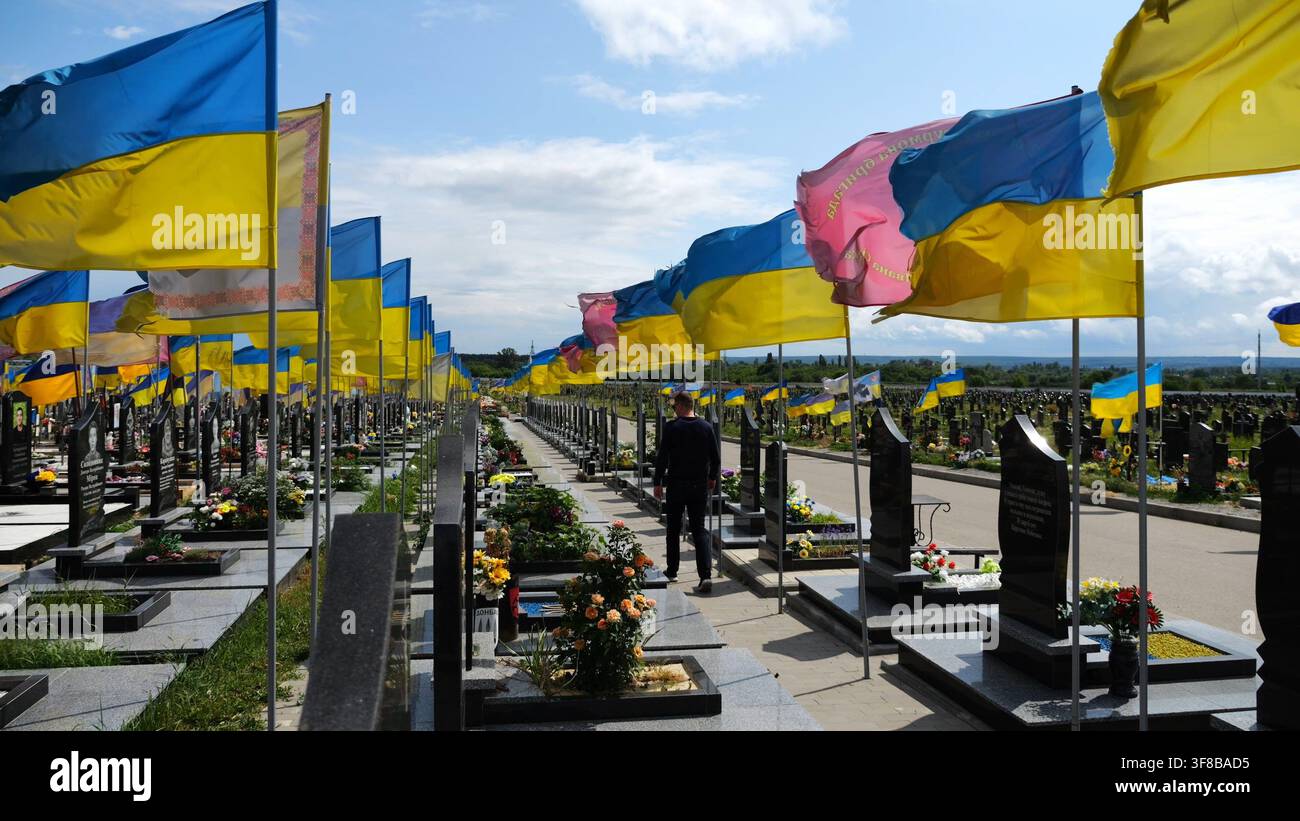 L'uomo in lutto va tra le innumerevoli tombe di soldati ucraini con bandiere nazionali nel cimitero militare di Charkiv. La scena riflette il dolore personale e. Foto Stock