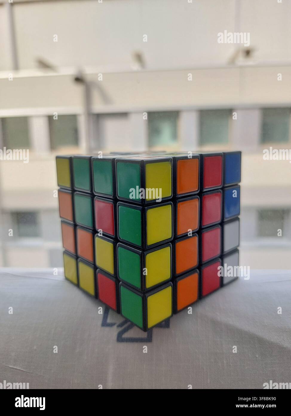 Prospetto frontale di un cubo di Rubik quattro per quattro posizionato su un foglio bianco accanto a una finestra. Foto Stock