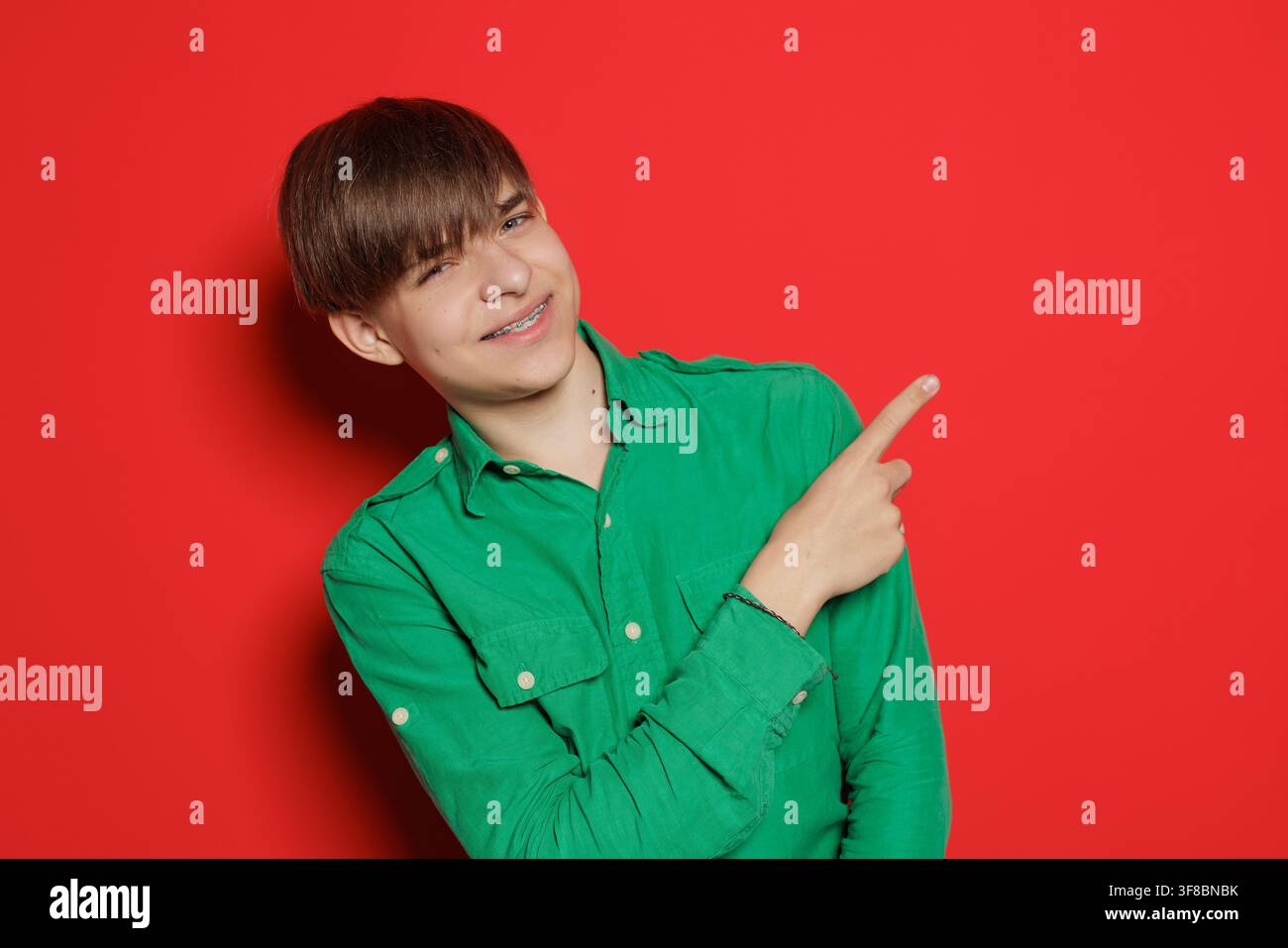 Un giovane uomo con una camicia verde sorride mentre indica di lato su uno sfondo rosso vivace. Foto Stock