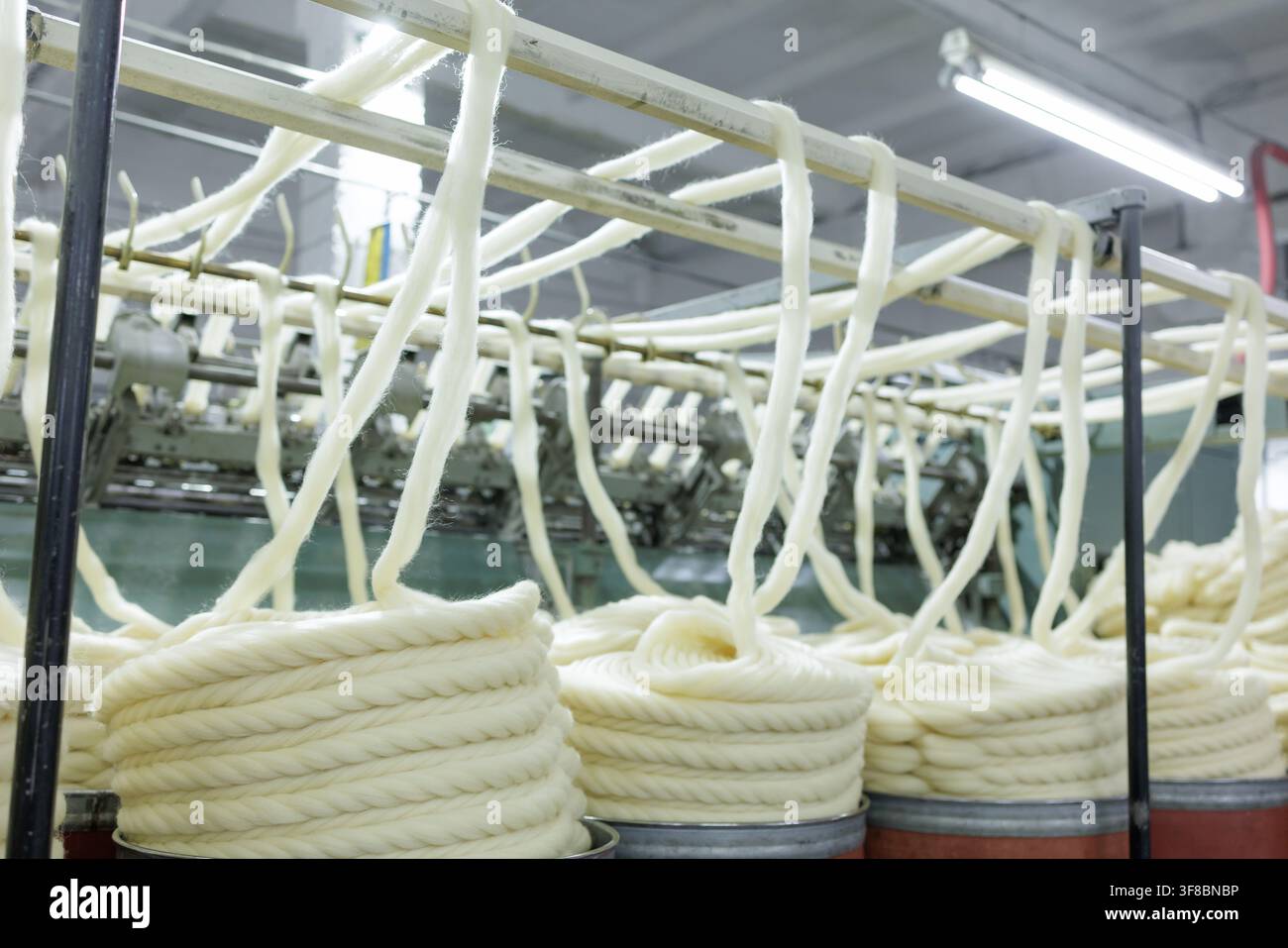 Spessi filoni di lana merino pendono da macchinari automatizzati e si avvolgono in bobine all'interno di una fabbrica tessile durante la filatura e la torcitura industriali Foto Stock