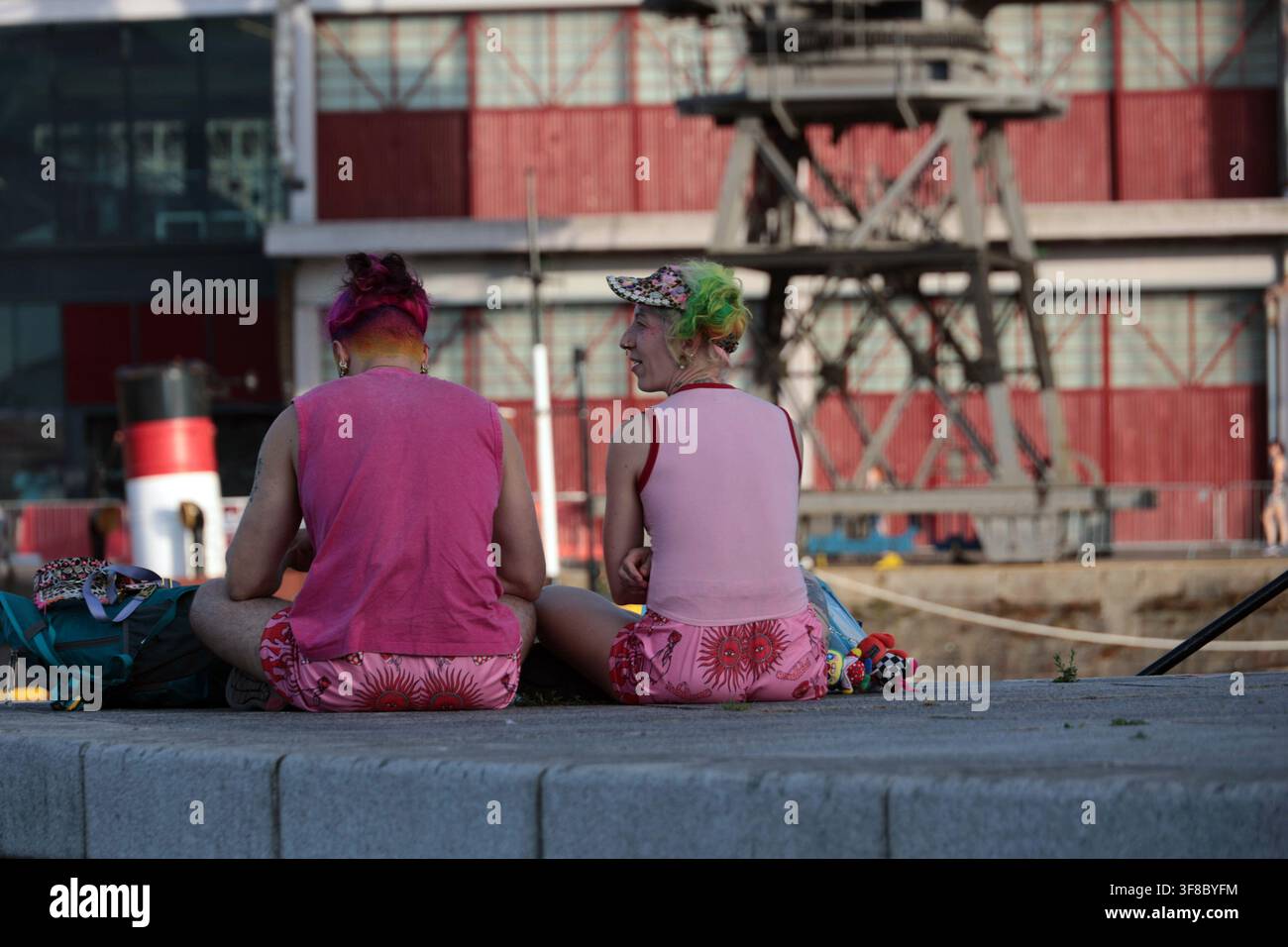 Stile di vita alternativo e sottocultura urbana: due persone con capelli colorati sedute casualmente vicino a un molo in un'area portuale industriale. Individualità e sé Foto Stock