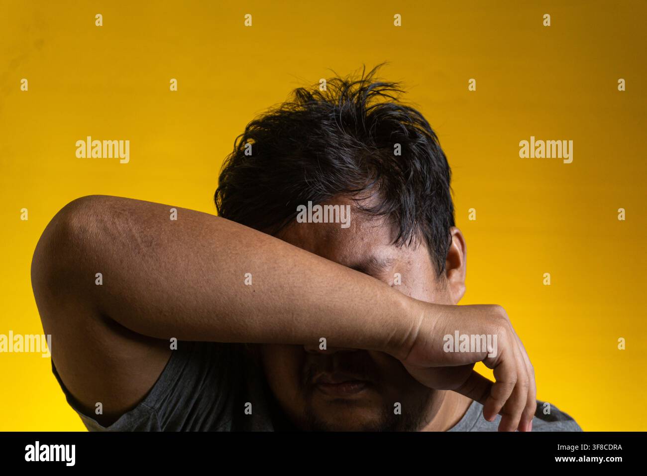 Un ritratto emotivo di un giovane uomo asiatico che nasconde il viso seppellendolo nella curva del braccio piegato. Il soggetto sembra sopraffatto, esausto Foto Stock