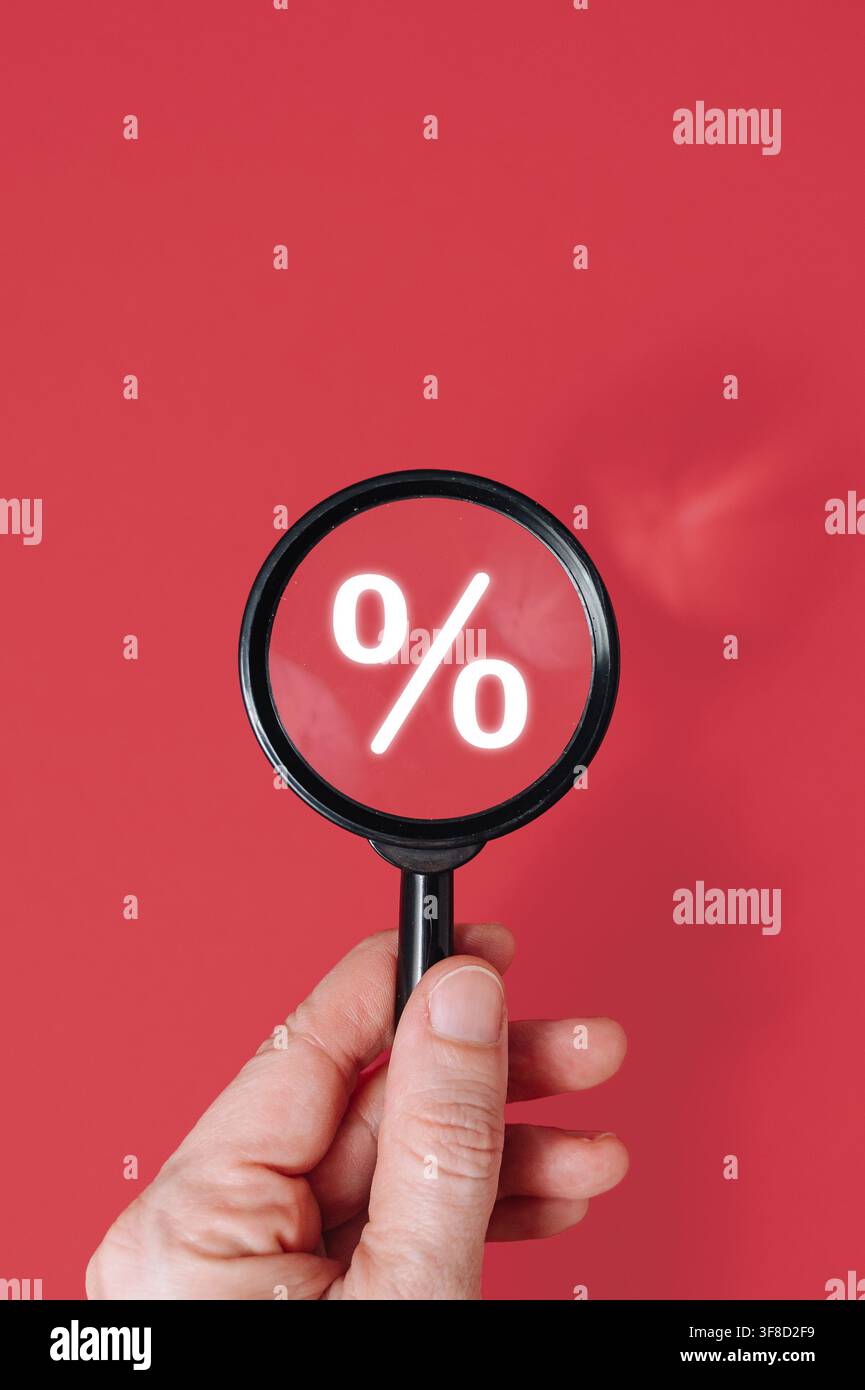 Mano che tiene una lente d'ingrandimento sopra un'icona di percentuale su sfondo rosso, ricerca di sconti caldi e concetto di vendita finale. Foto di alta qualità Foto Stock