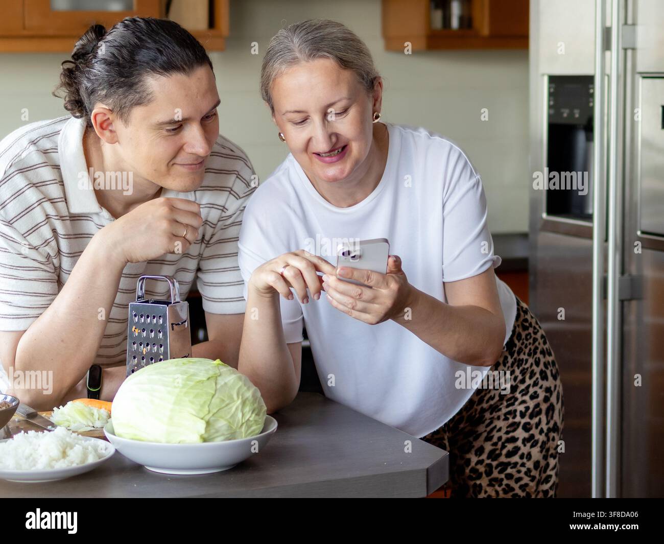Coppia sorride mentre controlla una ricetta sullo smartphone accanto agli ingredienti per gli involtini di cavolo fatti in casa. Concetto di tempo di cottura condiviso, connessione familiare, foo Foto Stock