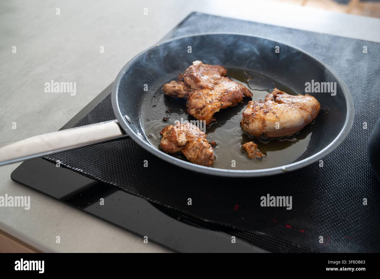 Cosce di pollo condite che sfrigolano nella padella su un piano cottura a induzione nero con vapore che sale, crosta dorata, spatola di metallo e luce naturale da Foto Stock