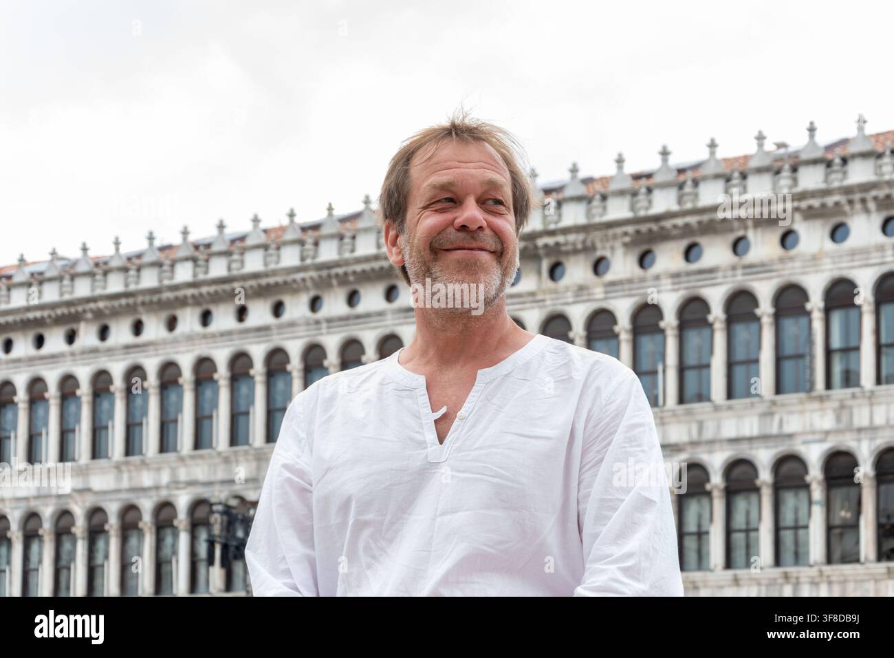 Un uomo maturo sorridente con una camicia bianca si trova all'aperto davanti a un edificio ad arco storico sotto un cielo nuvoloso. Foto Stock