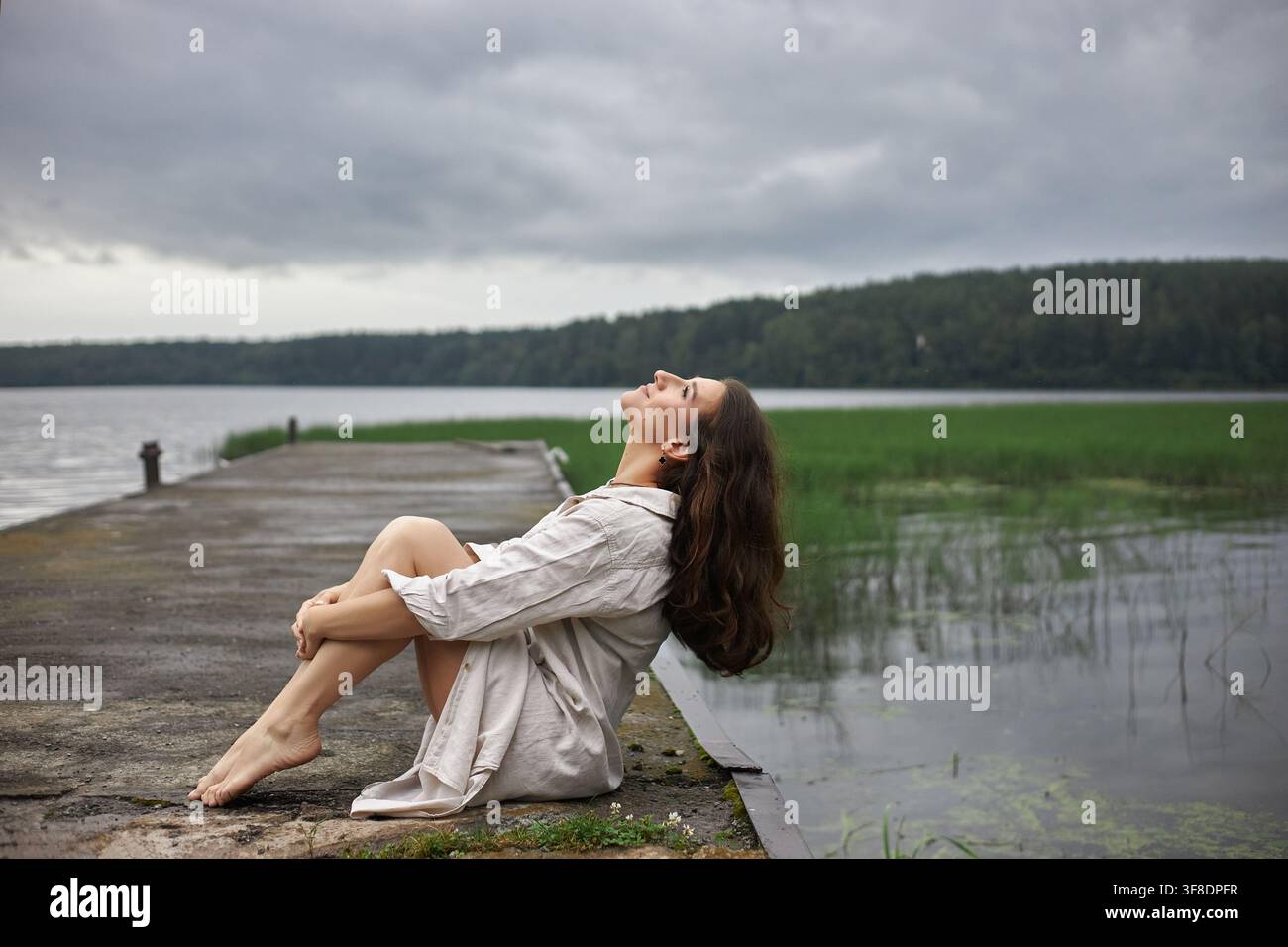 Donna seduta su un molo vicino a un lago con erba nell'acqua. È a piedi nudi e si appoggia all'indietro con gli occhi chiusi. Le nuvole riempiono il cielo, indicando un potenziale Foto Stock