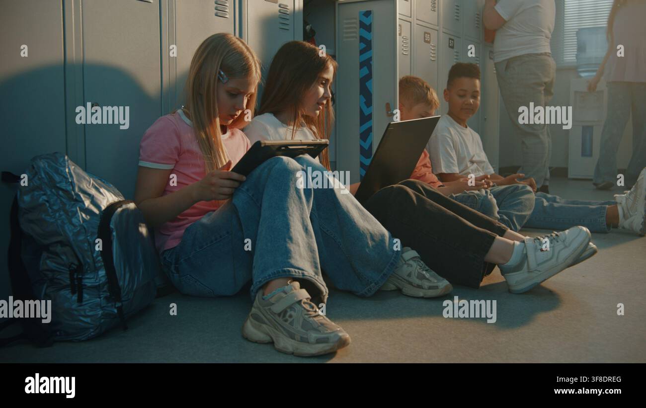 Due scolarette sedute per terra vicino agli armadietti, lavorano insieme su un laptop e un tablet digitale durante la ricreazione. Un ragazzo è seduto nelle vicinanze, concentrato sul suo smartphone. Alfabetizzazione digitale, tecnologia educativa moderna. Foto Stock