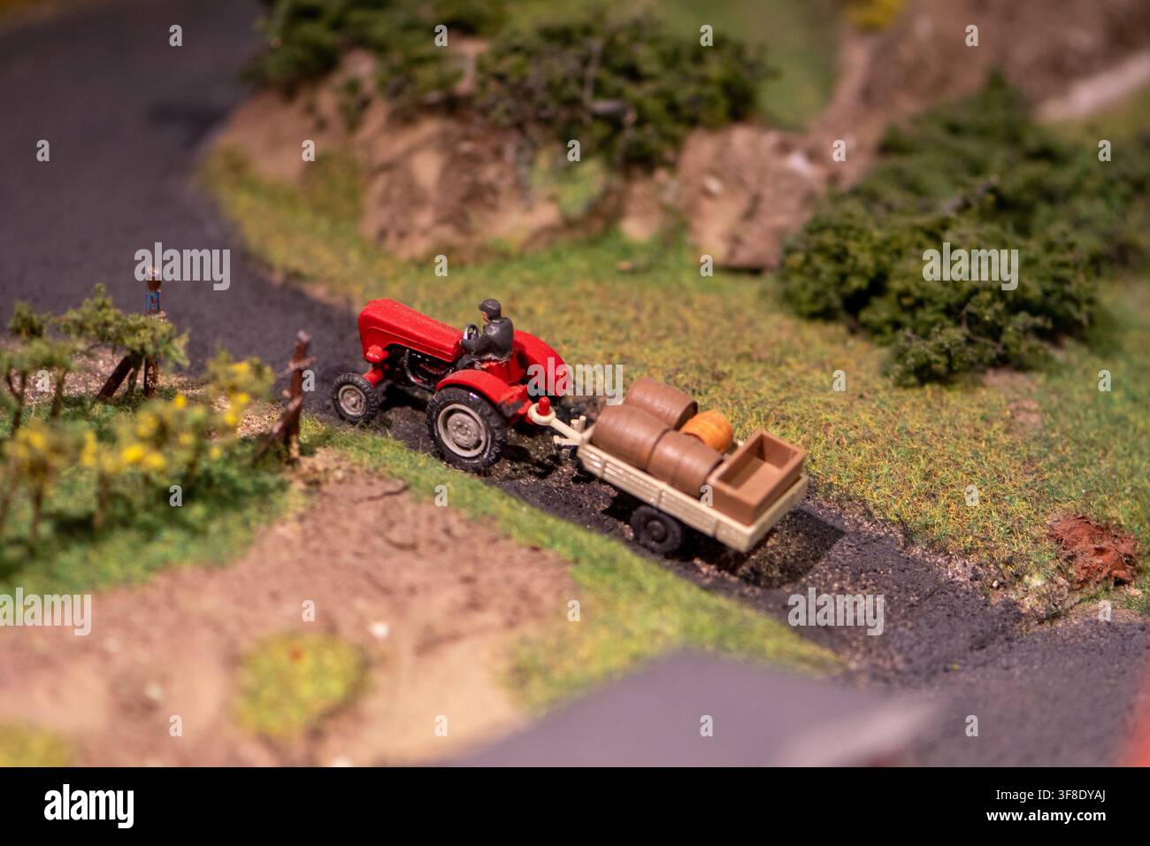 Un trattore rosso in miniatura con un piccolo rimorchio che trasporta barili si muove lungo una strada stretta in una scena di diorama rurale dettagliata. Foto Stock
